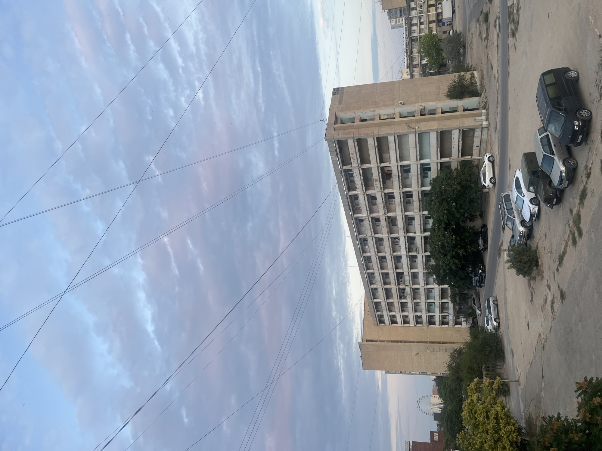 The image captures a view of a residential building in Aktau, Kazakhstan, under a cloudy sky. The building, a long, multi-story structure with balconies, appears to be made of concrete and exhibits signs of aging. Numerous power lines crisscross the sky above the building. In the foreground, several parked cars are visible on a dusty, paved area. The background includes trees, other buildings, and a Ferris wheel in the distance, suggesting an urban environment. The lighting and cloud formations indicate it is either early morning or late afternoon.