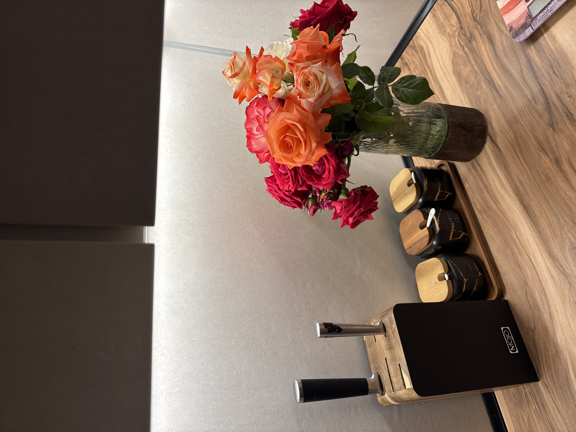 An indoor still life photograph showcases a kitchen counter with various items. On the left, a wooden knife block holds two knives. To the right of the knives, three spice jars with wooden lids are arranged on a wooden tray. In the center right, a bouquet of colorful roses in various shades of red, orange, and white is displayed in a clear glass vase with a textured base. The counter is made of wood with a visible grain pattern. In the background, a textured wall is illuminated by strip lighting from above, with dark cabinets overhead. The scene appears to be in a domestic kitchen setting in Mikhaylovsk, Russia.
