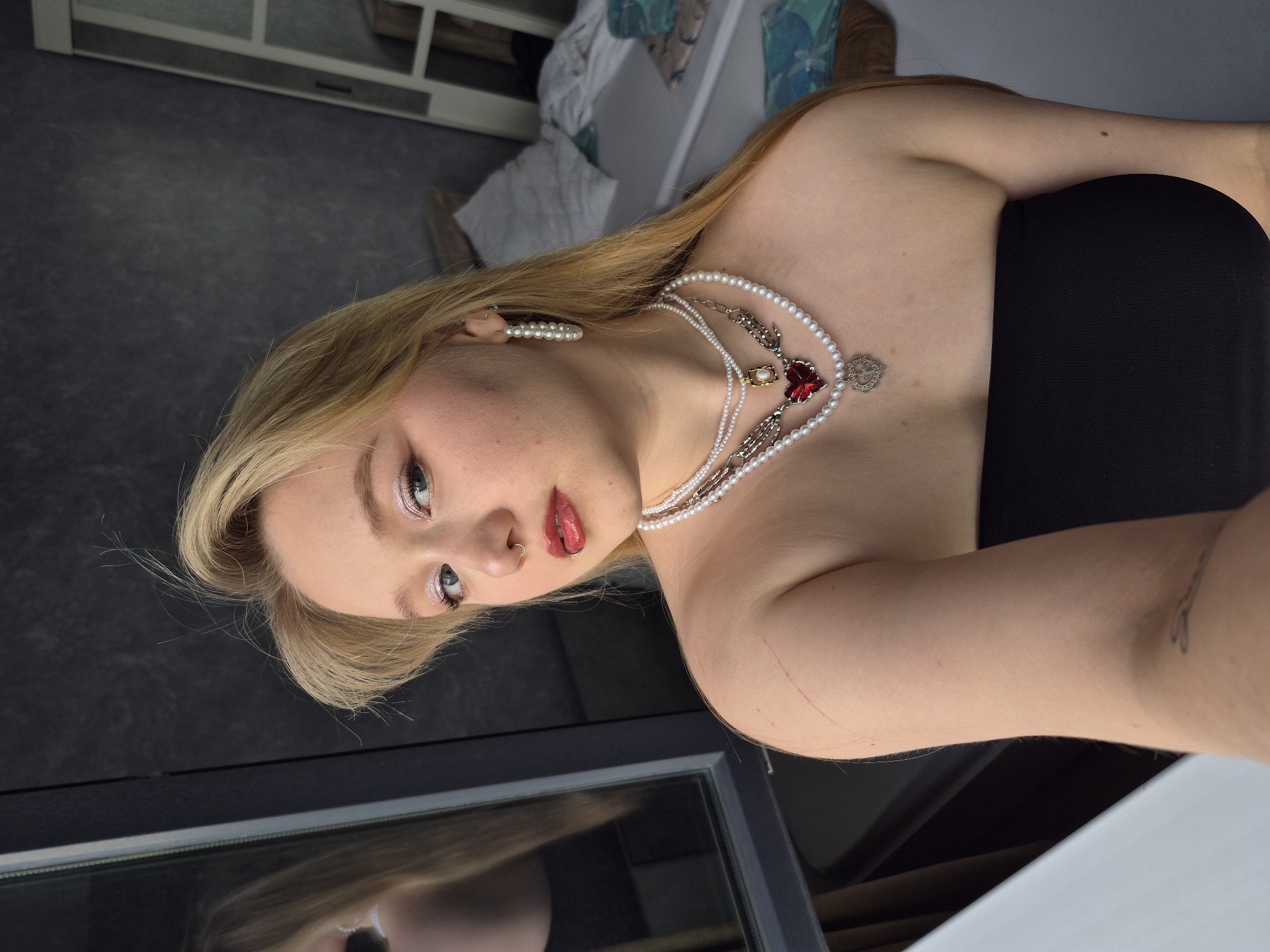 A young Caucasian woman with blonde hair and blue eyes is taking a selfie indoors. She is wearing a black strapless top and multiple pearl necklaces, one of which has a red heart-shaped pendant. She also has hoop earrings and a nose ring. Her makeup includes pink eyeshadow and red lipstick. There are visible scratches on her right shoulder and a small tattoo on her left arm. The background shows a bed with crumpled sheets, a window with a reflection of the woman, and a closet. The setting appears to be a residential bedroom. The woman's posture and the indoor environment suggest a casual, personal moment captured in a private space. The image does not contain any specific indicators of its location within the city of Izhevsk, Russia.