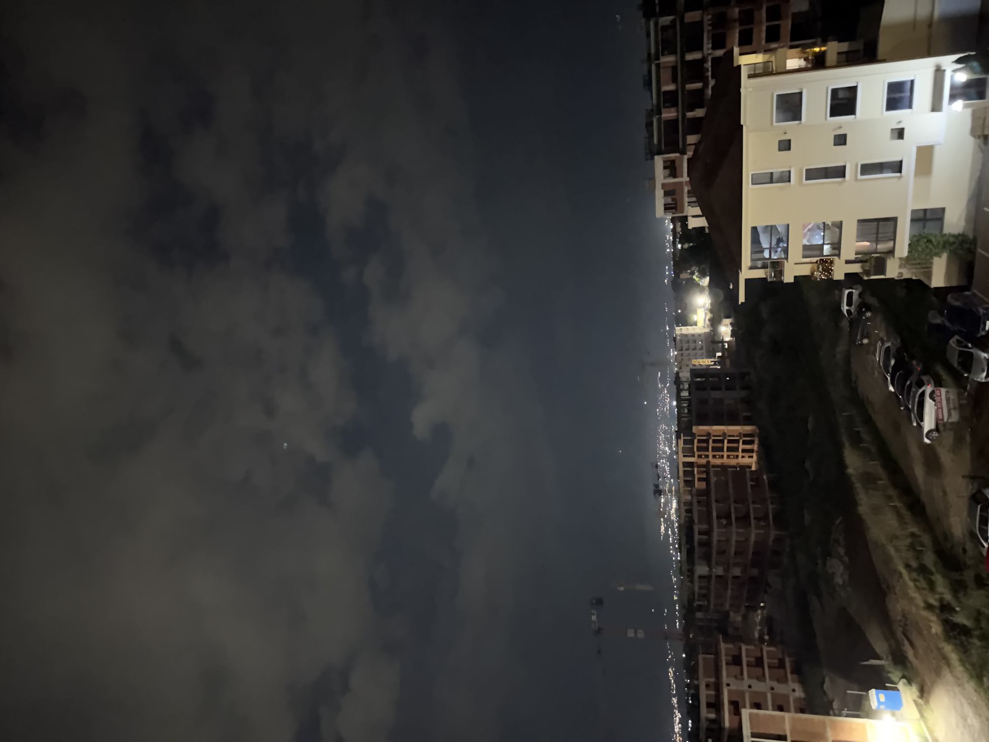 The image presents a panoramic nighttime view of Plovdiv, Bulgaria, seen from an elevated perspective. The scene is broadly divided into three horizontal segments when viewed in its correct, unrotated orientation.
The uppermost portion of the image shows a dark, cloudy night sky. Patches of lighter grey clouds are interspersed across the dark expanse, indicating an overcast atmosphere. A very faint, small bright dot, possibly a distant star or light source, is subtly visible within the clouds.
The middle section captures the vast expanse of the city, with a prominent horizon line of artificial lights stretching across the frame. Numerous small, bright points of light, predominantly yellow and white, illuminate the urban landscape, indicating buildings and streetlights that extend far into the distance. Several dimly lit, tall structures, consistent with construction cranes, are silhouetted against the distant city glow, suggesting active development.
The bottom section of the image constitutes the immediate foreground. On the right, a multi-story building with a light-colored (beige/cream) facade is prominent, with several of its windows illuminated. To its left, the incomplete skeletal structures of another large building suggest an ongoing construction project. Below these buildings, an unpaved or dirt area serves as a parking lot, filled with several parked cars of various models and colors, including a white SUV and a partially visible red car. A small blue portable toilet is positioned near the bottom left corner of this area. Sparse greenery and foliage are also visible around the structures and along the ground.
The overall impression is that of a quiet urban night, with the artificial lights of the city providing the primary illumination under a cloudy sky. No specific human activity or event is distinctly captured. There is no clearly legible text visible within the image.