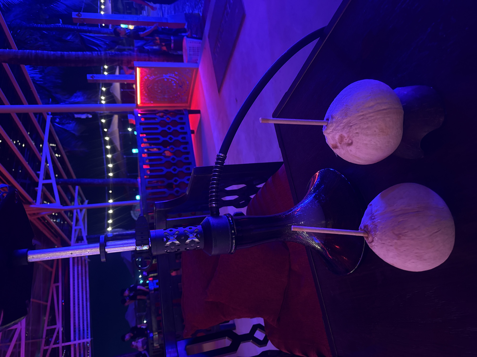 Here is a description of the image:
The image shows a hookah with two coconut halves pierced with sticks in place of a traditional mouthpiece and bowl. The hookah is black with a long, flexible hose and sits on a dark wooden table. Red pillows are visible behind the base. The background features an outdoor setting with string lights, palm trees, and architectural elements with red and blue lighting, suggesting a bar or lounge atmosphere at night. The image is taken from a close-up, angled perspective. The location is Unknown, Unknown.