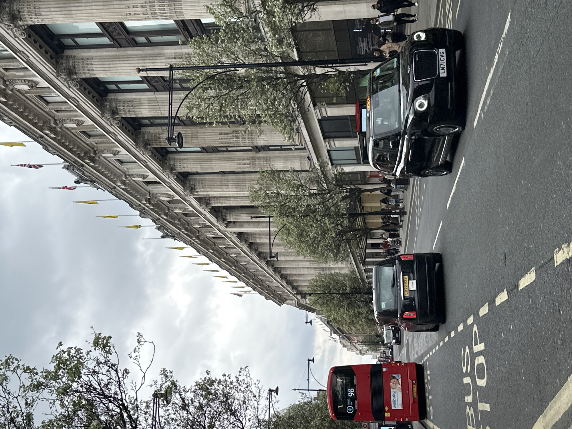 The image depicts a street scene in London, United Kingdom. A grand building lines the left side of the street, adorned with classical architectural details such as columns and flags. Several people are visible along the sidewalk next to the building. A red double-decker bus and a black taxi are prominent on the right side of the road. The street is marked as a bus stop. The sky is overcast, and trees with budding leaves are visible at the lower left, contributing to the urban landscape.
