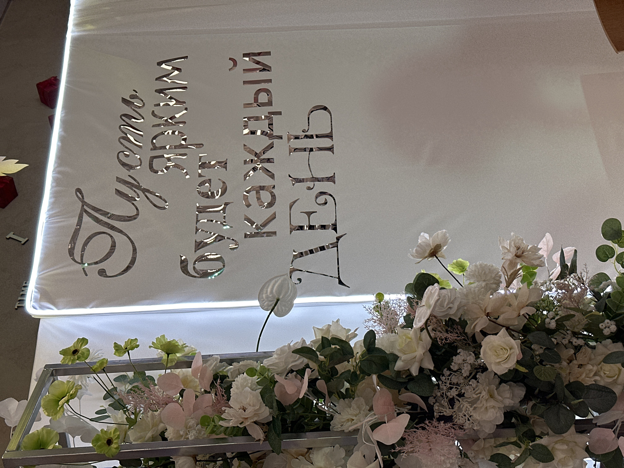 Here is a description of the image:
The image shows a decorated wedding reception or event in Chaikovsky, Russia. A white panel with silver lettering in Russian is visible, reading "Пусть что будет ярким каждый день" (May every day be bright). The panel has built-in lighting along its edges. The panel is decorated with artificial flowers, predominantly white and pale pink, set within a gray metallic frame. The floral arrangement includes roses, leafy foliage, and other decorative plant elements. The scene is well-lit and appears to be indoors.