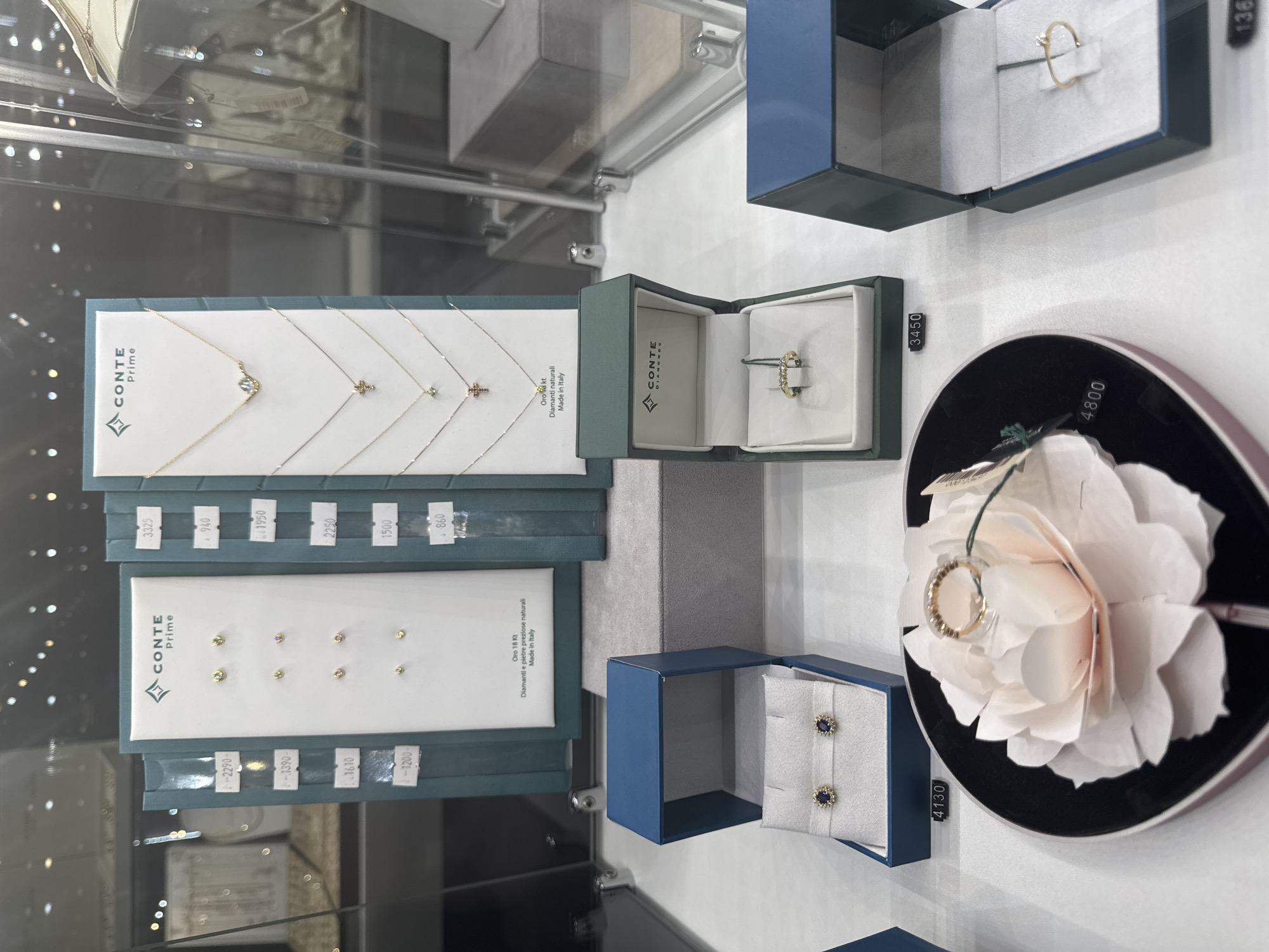 The image shows a jewelry store display in Batumi, Georgia, featuring various gold jewelry pieces, including earrings, necklaces, and rings. The display is organized and well-lit, with each item presented in a box or on a stand. Some items have price tags associated with them, such as "4130", "3450", and "4800". The jewelry is primarily gold with diamonds and colored gemstones, suggesting a high-end establishment. The store appears to be a modern retail environment with glass display cases. There are no people visible in the image. The overall impression is one of a sophisticated jewelry store showcasing its merchandise.