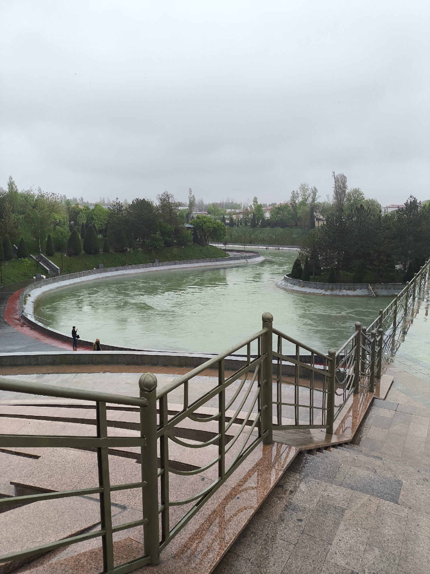 The image depicts a grey and overcast day at the Ankhor Canal in Tashkent, Uzbekistan. A wide canal or artificial lake, with a greenish-grey water surface, occupies the central part of the scene. The canal has curved edges and is bordered by paved walkways, grassy areas, and trees. A man and a woman are standing near the edge of the canal. A metallic green railing runs along the right side of the image, leading down a set of stone steps. The ground is wet, and reflections of the railing are visible on the wet surfaces. The background features trees and low-rise buildings, obscured by the overcast sky.