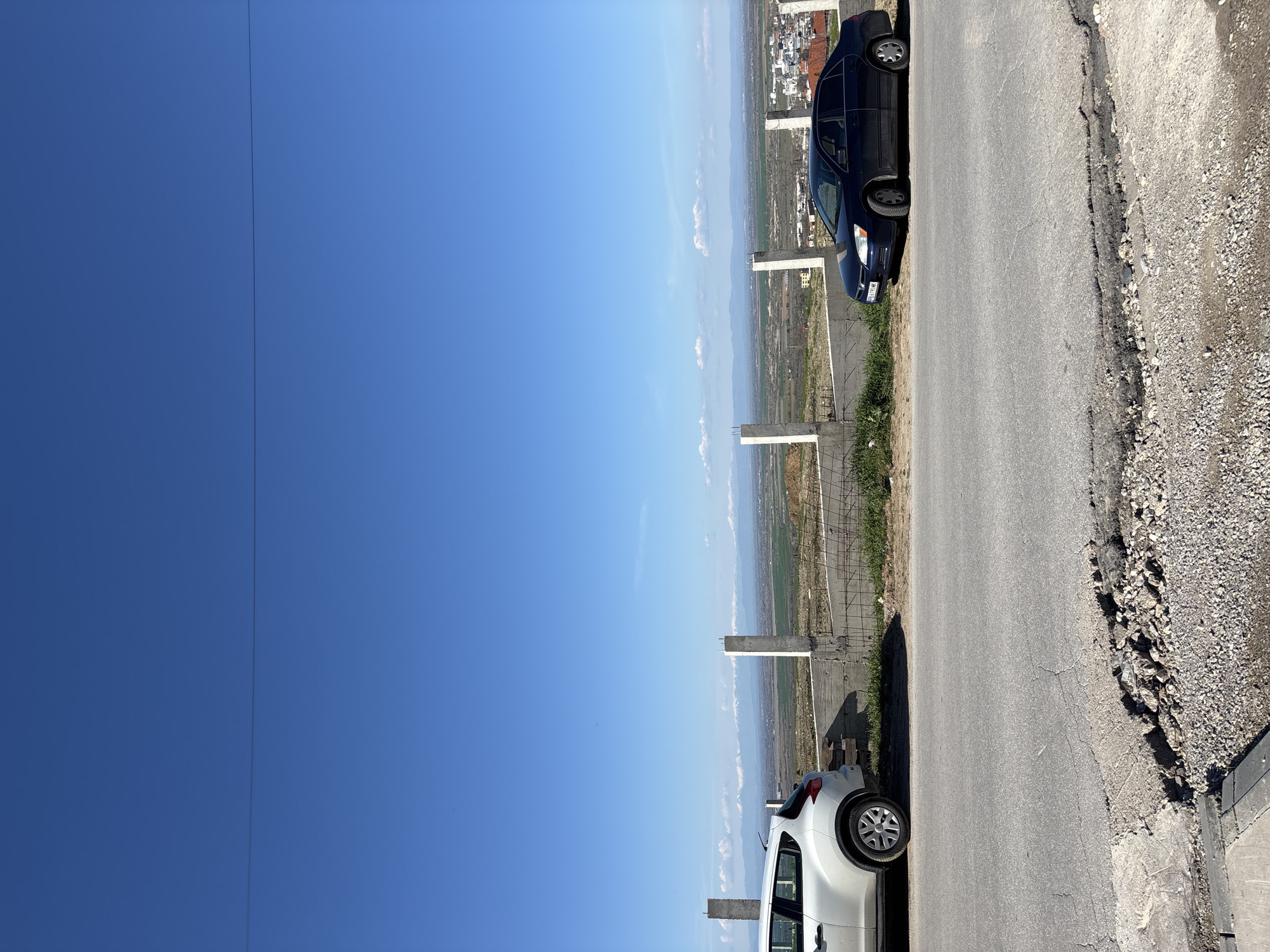 The image depicts an outdoor scene on a clear day with a bright blue sky. A road runs along the right side of the image, showing signs of wear and tear with a rough shoulder area of loose gravel. Parked on the road are two cars: a white vehicle and a dark-colored vehicle, both situated along the edge of the road. A fence constructed from concrete poles runs parallel to the road, separating the road from a distant view of land, possibly a valley or field, with traces of a city in the far background. The exact location is Asenovgrad, Bulgaria.