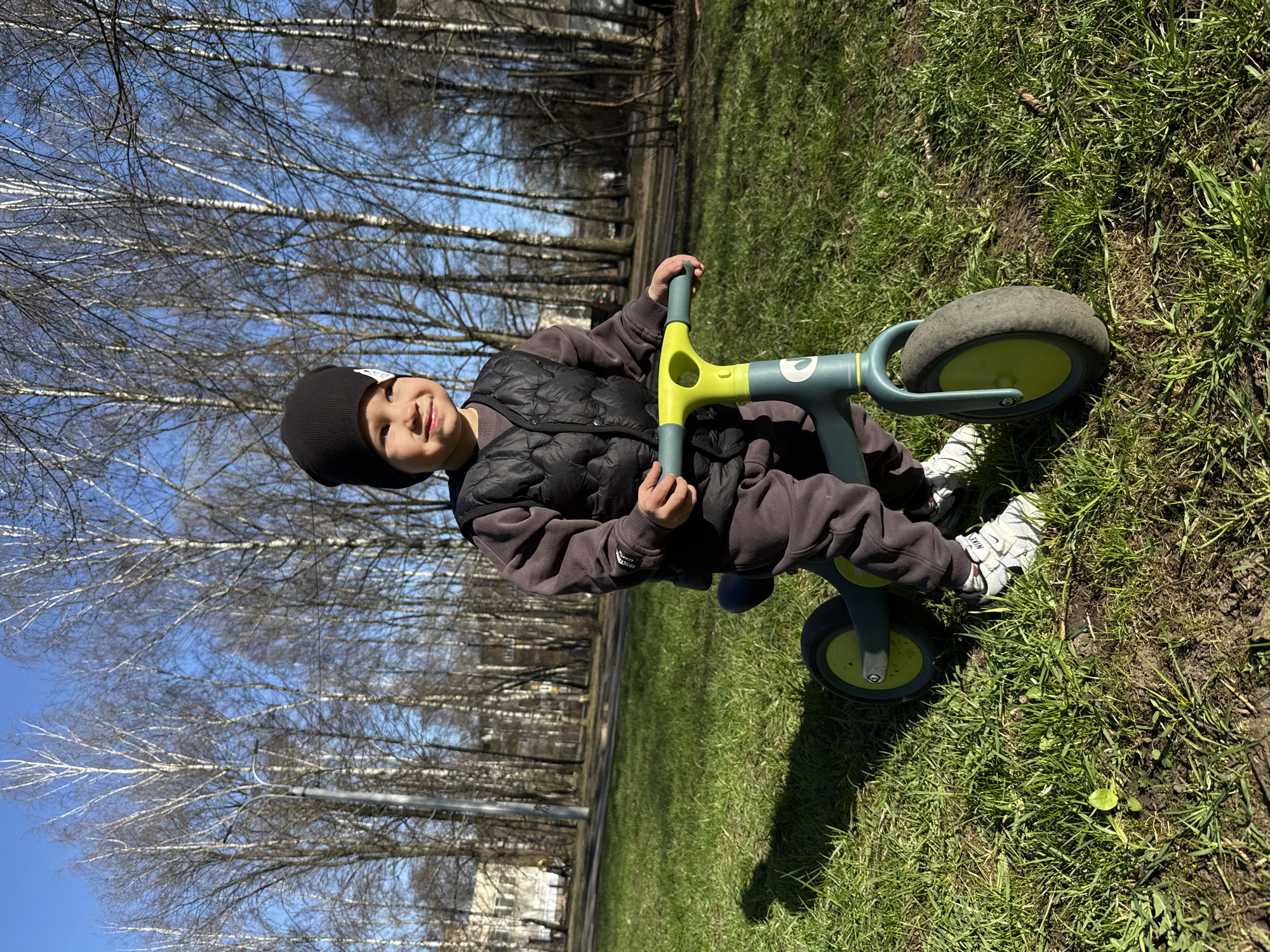 A young child, wearing a black beanie, a dark quilted vest, and grey athletic pants, sits on a green and grey balance bike in an outdoor setting. The child is smiling and holds onto the bike's handlebars. The bike rests on green grass. Bare trees, likely birch, are visible in the background against a blue sky. Based on the architecture and the trees, the location is likely in Rusanovo, Russia. There's a building visible behind the tree line, with a street light visible. The scene appears to be taken on a sunny day with sparse, early spring vegetation.