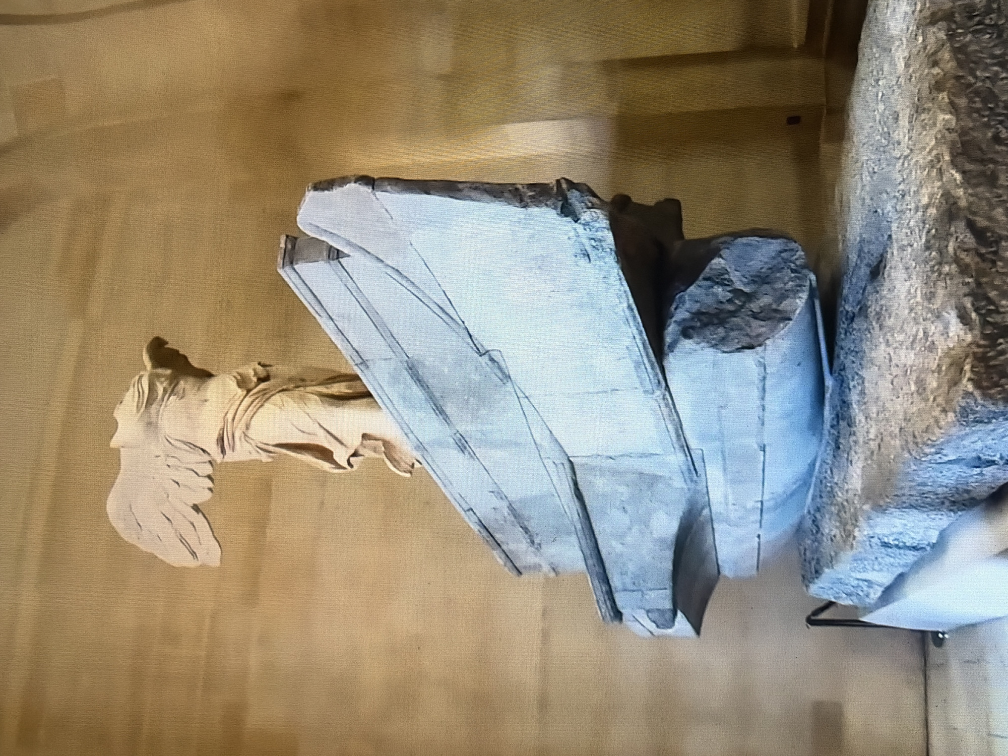 A headless, winged statue of Nike stands atop a layered stone pedestal within a museum. The statue is made of light-colored stone and is draped in a garment. The pedestal is composed of several large, irregularly shaped blocks of grey stone. The background is a muted tan wall. The statue is likely the Winged Victory of Samothrace, a famous Hellenistic sculpture.