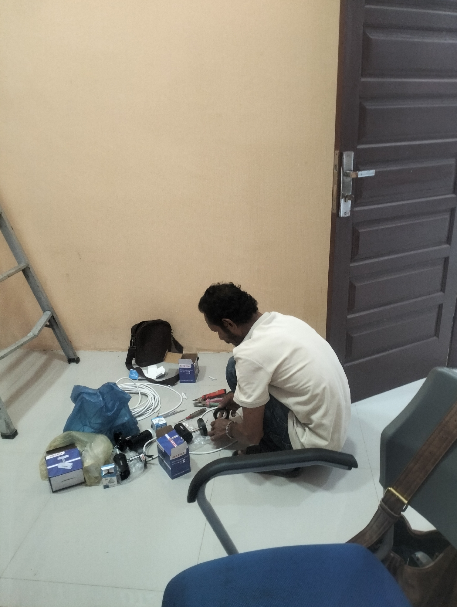A person is kneeling on the floor of a room, working on electrical wiring. There are several boxes of electrical components, coiled white wire, a blue plastic bag, and tools scattered around the person. A ladder is leaning against the wall to the left, and a dark brown door is visible on the right. The walls are painted a light tan color, and the floor is tiled. The person is wearing a light-colored t-shirt and dark pants. The scene appears to be in an indoor setting, possibly an office or a residential building, in the city of Lhokseumawe, Indonesia.