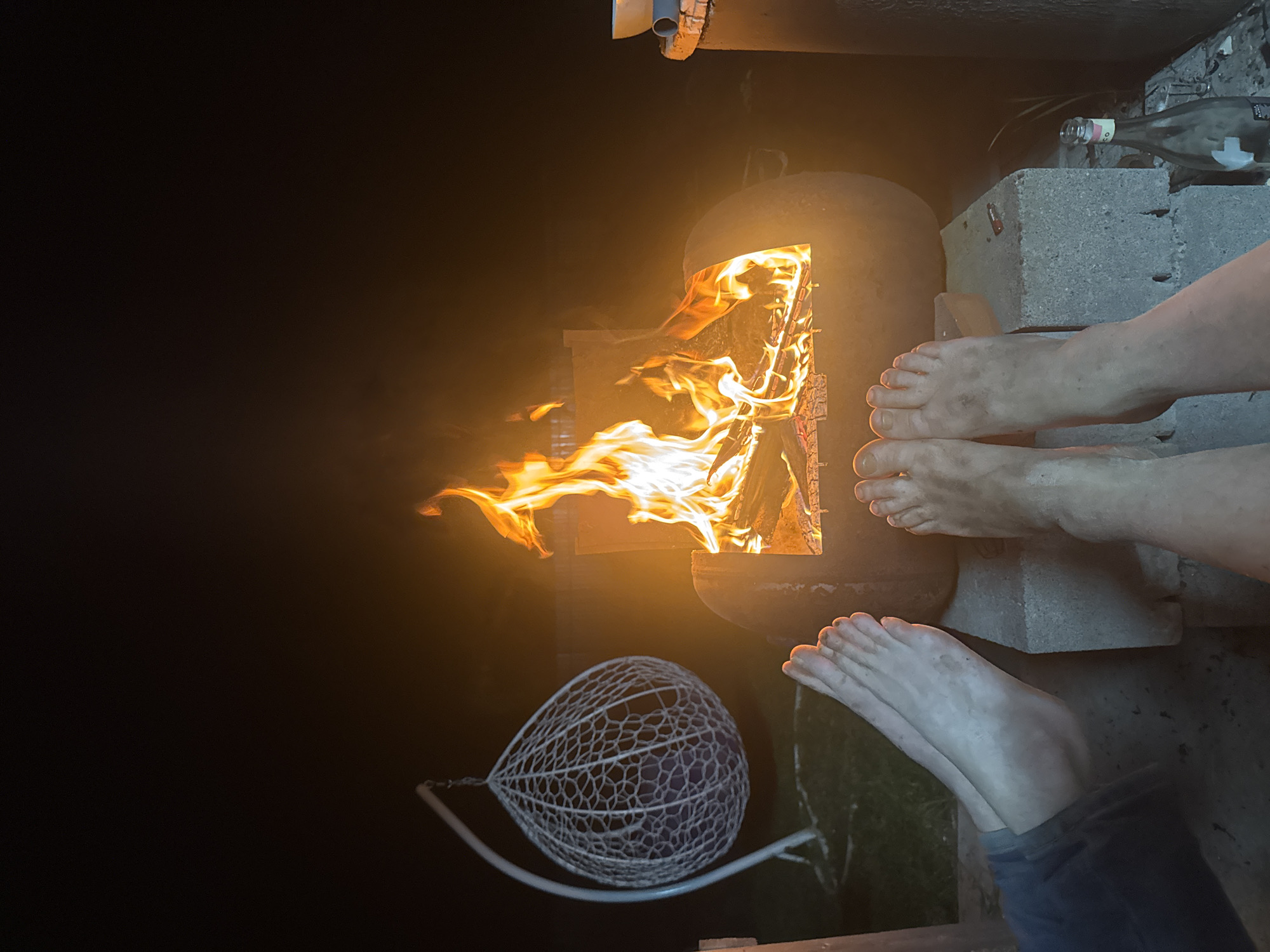 The image depicts an outdoor scene at night, likely in a backyard setting. A makeshift fire pit constructed from a metal tank or drum with a rectangular opening serves as the focal point. Flames and burning wood are visible inside the fire pit, casting a warm glow on the surroundings.
To the right of the fire pit, two pairs of bare feet are positioned near the fire, suggesting that people are warming themselves. The feet are of varying sizes and skin tones, indicating they belong to different individuals. One pair of feet is resting on concrete blocks, which also support a discarded glass bottle.
To the left of the fire pit, partially obscured by darkness, is a white netting hammock chair.
The image lacks specific details to confirm its location within the city of Chaikovsky, Russia.