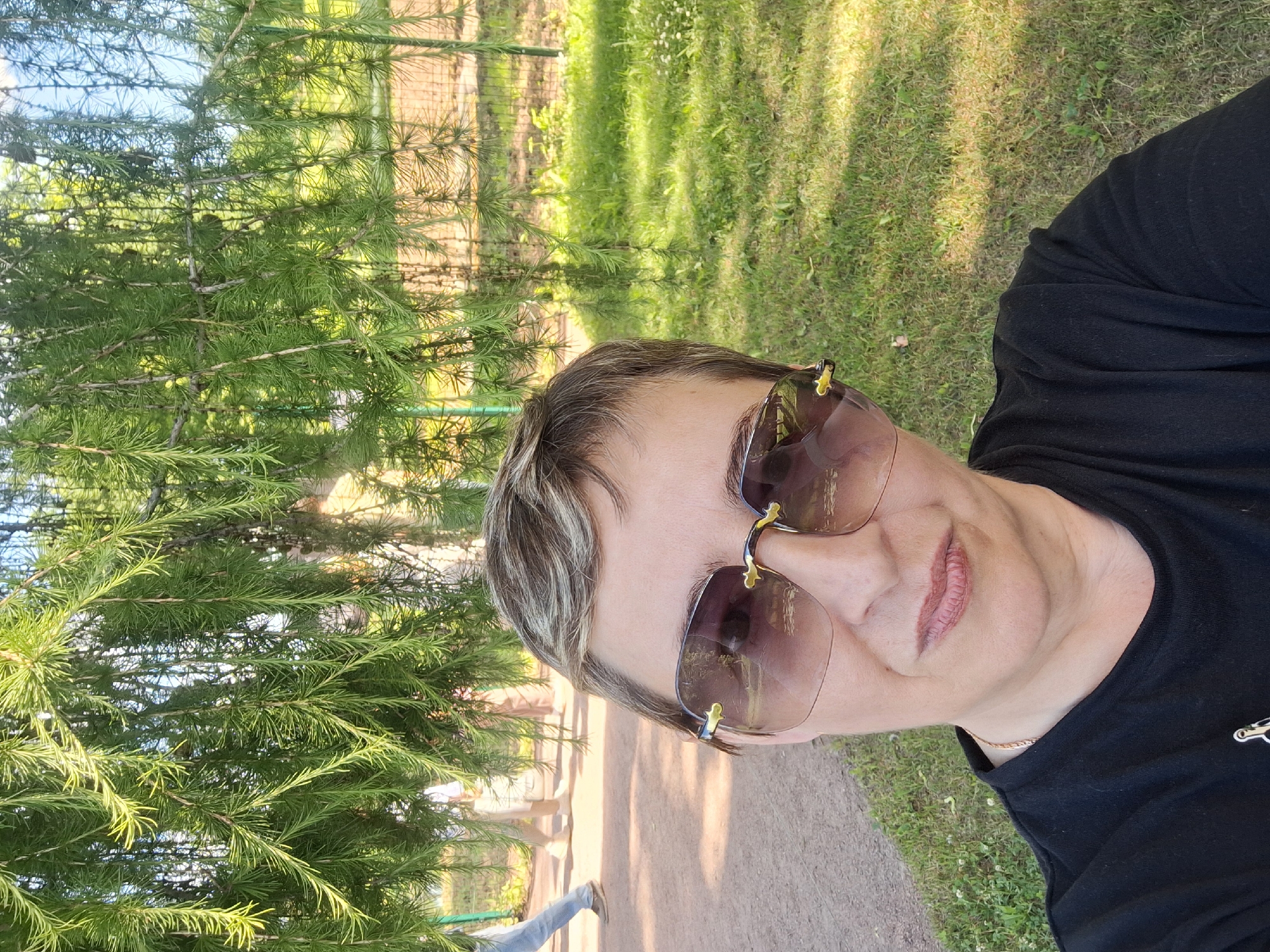 A woman with short, light-brown hair and blonde highlights, wearing large, rimless sunglasses with brown lenses and gold accents, and a black t-shirt, takes a selfie outdoors. She is smiling slightly. Behind her, a pathway is visible, with two people walking in the background. The environment is green and sunny, with grass and trees, suggesting a park setting. The scene is likely in Gatchina, Russia, possibly near the Gatchina Palace or a park within the city.