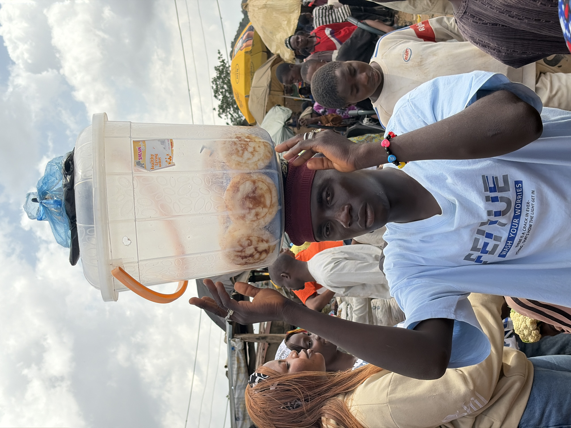 In Gura Top, Nigeria, a young Black male with a maroon kufi hat precariously balances a large, clear plastic tub filled with what appear to be round, baked goods on his head. He is holding the tub steady with both hands. The tub has an orange handle on one side and a blue plastic bag is tied to the top. A label on the tub reads "SALVIN Healthy Living Tucker". The man is wearing a light blue t-shirt with a graphic that reads "FEIUE WASH YOUR WORRIES THERE IS A CRACK IN EVER- THING, THAT IS HOW THE LIGHT GET IN". He is also wearing a colorful beaded bracelet on his right wrist. In the background, a busy market scene unfolds with other individuals, market stalls, and umbrellas providing shade. A woman with long reddish-brown hair and a tan hooded sweatshirt is visible to the left, looking towards the man. The sky above is overcast with clouds.