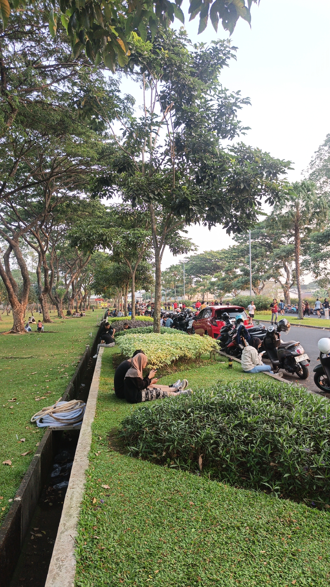 The image depicts a park scene in Bacin, Indonesia, with people relaxing on a grassy area. In the foreground, two individuals are seated on the grass, one wearing a hijab and both looking at their phones. To their left, a concrete drainage ditch runs along the edge of the lawn, containing a coiled blue hose. Behind the seated individuals, a variety of parked motorcycles and a red car are visible, suggesting a public gathering or popular recreational spot. The background features a treelined street with more people walking and standing. The environment is characterized by lush green grass, mature trees with full canopies, and a clear sky. The overall impression is of a relaxed outdoor atmosphere, possibly during a weekend or evening.