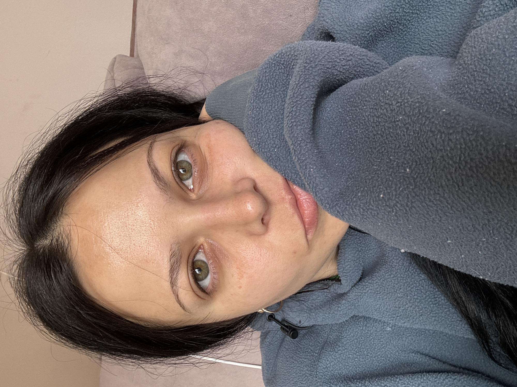A woman with dark hair is lying on a soft, beige-colored surface, likely a couch or bed. She is wearing a blue, fuzzy fleece sweatshirt with a drawstring. Her face is prominent in the frame, showing her green eyes, fair skin, and full lips. A gold hoop earring is visible on her left ear. The background is a plain, light-colored wall. The image doesn't reveal any information about the location in Pernik, Bulgaria.