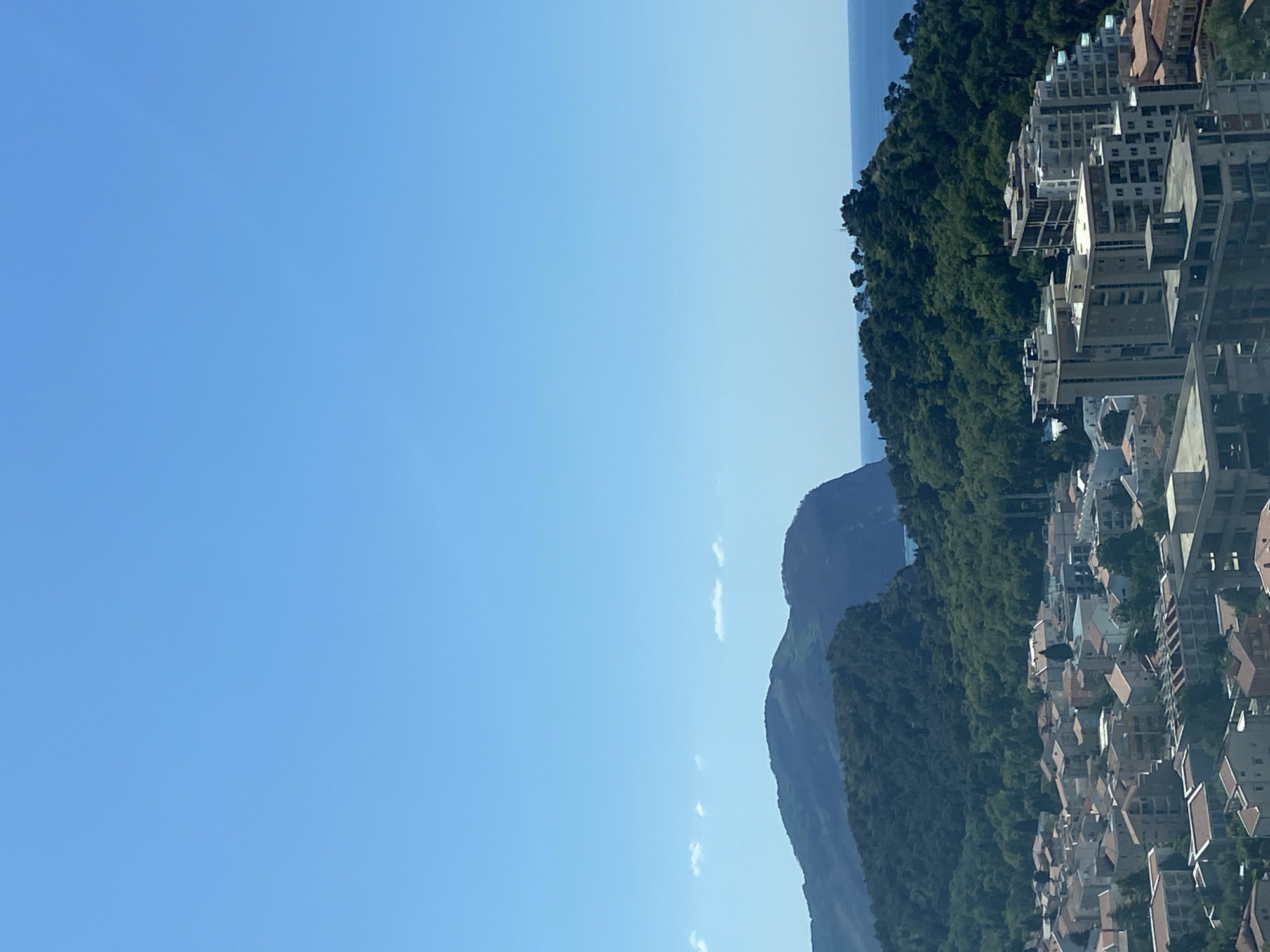The image presents a panoramic daytime view of a coastal landscape, consistent with the location context of Nerin, Montenegro. In the foreground and mid-ground, a dense cluster of urban buildings, likely residential and commercial, features predominantly light-colored facades and numerous terracotta-tiled roofs. These structures are built along a sloping terrain, suggesting a hillside settlement.
A prominent, densely forested mountain or large hill forms a natural barrier, extending horizontally across the mid-section of the frame, positioned behind the urban area. Beyond this forested expanse, the serene blue waters of the sea are visible on the horizon to the far right. Further to the left and receding into the distance, another, larger and more rugged mountain range with sparse vegetation can be seen, its peaks softened by atmospheric haze.
The sky is a clear, bright blue, indicating sunny weather and good visibility. The strong, even light suggests the time of day is either morning or early afternoon. No individuals, specific activities, or visible text are present in the scene, which conveys a sense of peaceful natural beauty intertwined with human habitation.