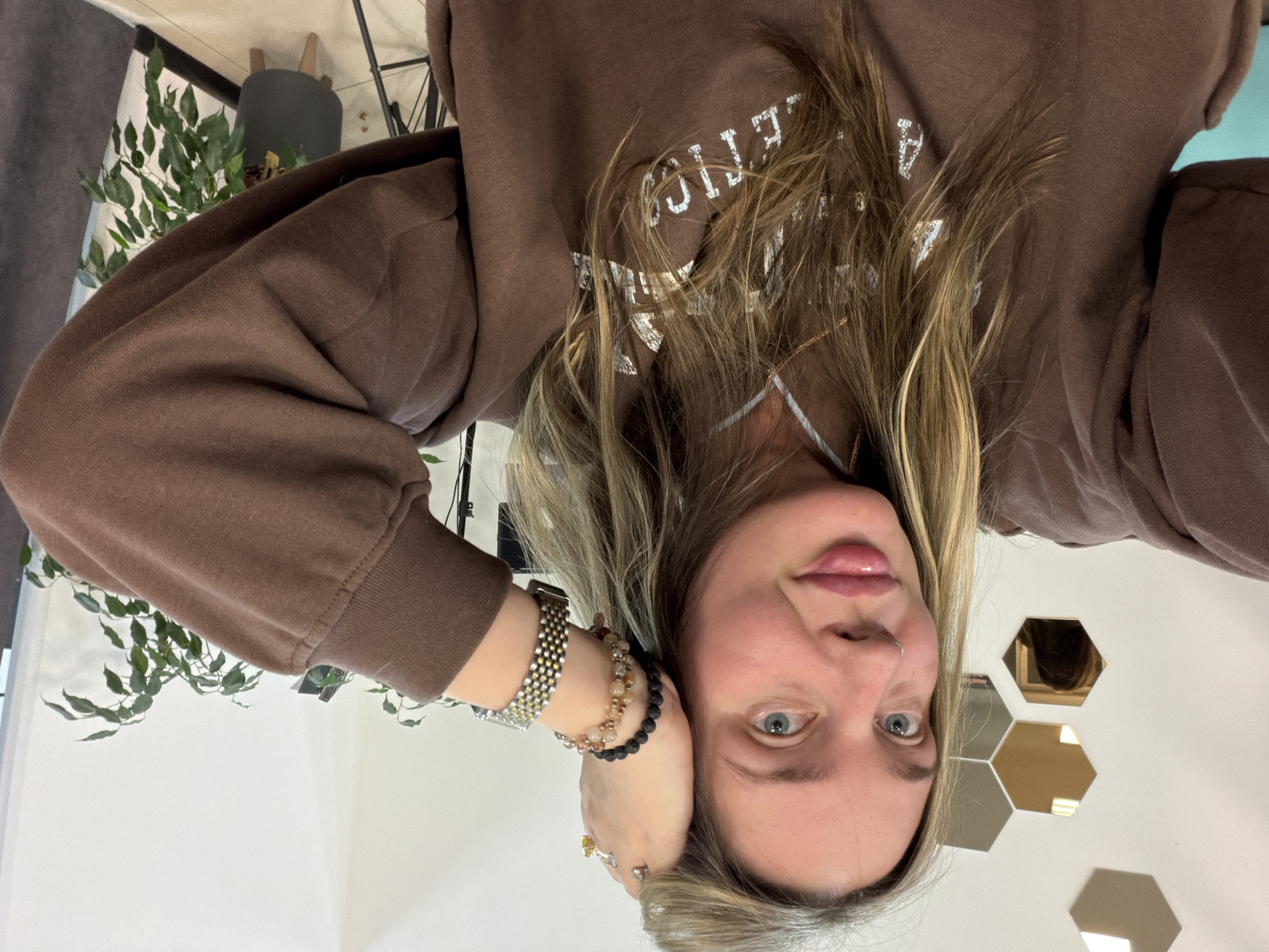 The image displays an upside-down portrait of a young woman. She looks directly at the camera with wide, light blue eyes and slightly parted lips, her expression appearing neutral to subtly engaged. Her long, light brown hair with blonde highlights is styled loosely, falling across her shoulders and chest.
She is wearing a brown long-sleeved sweatshirt or hoodie featuring white, collegiate-style text on the front. Visible parts of the text read "ATHLETIC" and "UNIVERSITY" or "COLLEGE". Her right arm is raised, with her hand resting near the side of her head. On her right wrist, she wears a silver and gold-toned watch alongside several beaded bracelets, including one made of black lava stones and others with lighter-colored beads. A ring with a yellow stone is visible on her right ring finger, and she has a small stud piercing in her right nostril.
The background indicates an indoor setting. To her left, a leafy green plant, possibly a Ficus, is partially visible, with a dark grey stool or chair and some metal framework behind it. To her right and behind her, a light-colored wall is adorned with multiple hexagonal panels, some of which appear to be reflective mirrors or decorative surfaces, showing indistinct reflections. The lighting in the room is bright and even. The scene suggests a casual indoor environment in Chaikovsky, Russia.