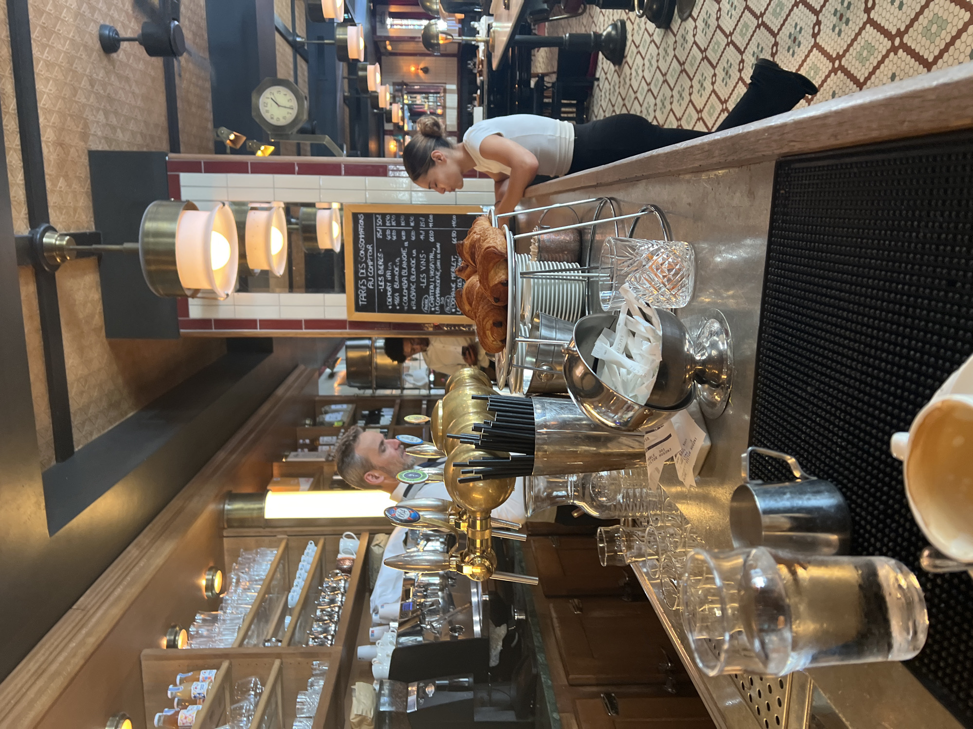 This is a medium shot, vertically oriented photograph of the interior of what appears to be a cafe or bar. The scene is bustling with activity. In the foreground, a man with short, graying hair and a beard is visible from the shoulders up, looking to his left. He is wearing a white shirt. He appears to be a bartender, positioned behind a polished brass beer tap system. Various glassware, including crystal tumblers and wine glasses, are neatly arranged on shelves behind him and on the counter.
On the counter, a basket of croissants sits next to a stack of small plates. A woman, with her back to the camera, is leaning over the counter, looking down at a chalkboard menu. She has her hair tied up in a bun and is wearing a white top and black pants. The chalkboard lists prices for drinks and food in French. Another person, a man, is visible further in the background, also looking towards the counter.
The overall setting suggests a warm, inviting atmosphere. The lighting is a mix of natural and artificial sources, casting a soft glow. The tiled floor and patterned wallpaper add to the establishment's decor. The presence of the menu board, beer taps, and glassware indicates that this is likely a place where drinks and possibly light food are being served. The time of day is not explicitly indicated, but the indoor lighting suggests it could be either day or evening. The mood appears to be relaxed and operational, with staff attending to customers and preparing orders.
Visible text includes a chalkboard menu with French text listing "TARIFS DES CONSOMMATIONS AU COMPTOIR" (Prices of drinks at the counter) and various drink and food items with their prices. There are also labels on bottles of beverages.