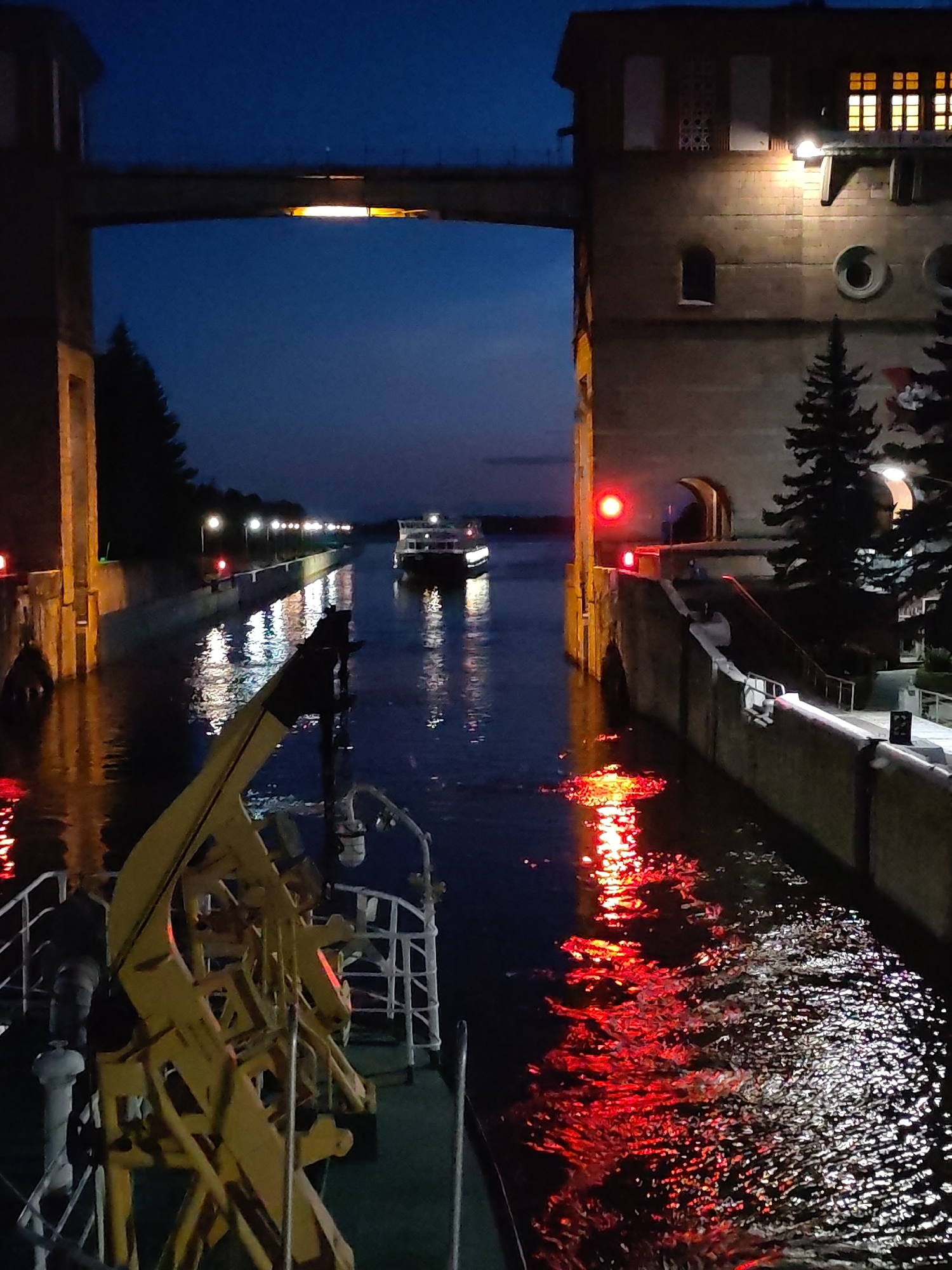 This is a nighttime photograph taken from the deck of a boat, looking towards a lock system at the city of Bol'shie Mkhi, Russia. The sky is a deep blue, transitioning to a darker hue at the top of the frame. A large, illuminated structure, possibly a building or part of the lock mechanism, stands on the right side of the image. It is made of stone or concrete and has several windows with warm, yellow light emanating from them. A horizontal bridge or walkway spans across the top of the frame, with a bright light fixture beneath it.
The water in the lock is dark and reflects the lights from the surrounding structures. There are red lights visible on the lock walls, indicating a signal or warning. In the distance, a white boat with lights is moving through the lock. Trees, silhouetted against the dark sky, are visible on the left bank of the canal. In the foreground, the deck of the boat from which the photo is taken is visible, with a large yellow mechanical arm or crane structure dominating the lower left portion of the image. The overall atmosphere is one of industrial infrastructure illuminated at night, with a sense of calm movement on the water.