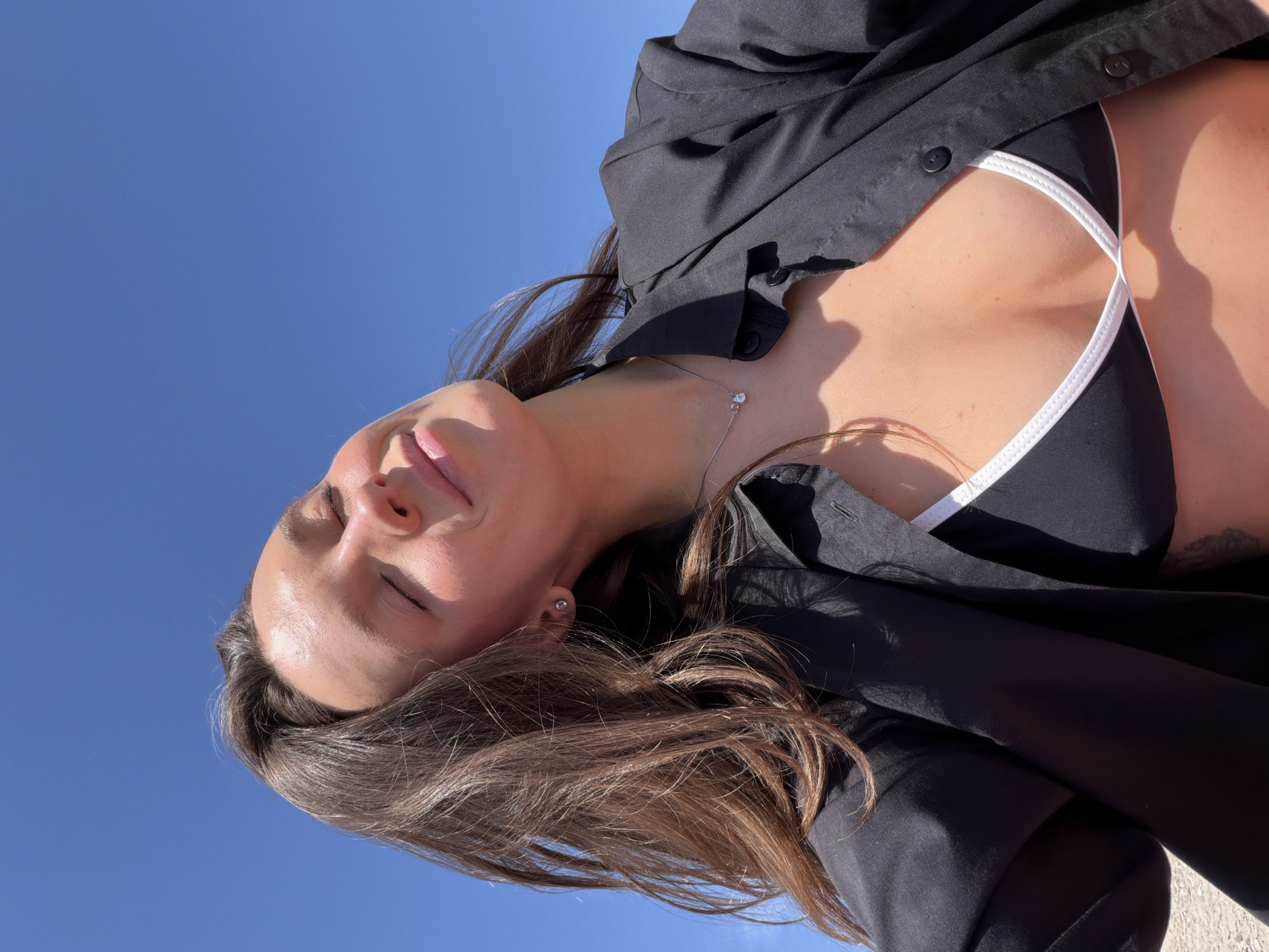 The image is a selfie of a woman in Milovići, Montenegro, taken from a low angle, looking up at her against a clear blue sky. She has long brown hair and is wearing a black shirt, which is unbuttoned to reveal a black top with white trim. She has her eyes closed and a serene expression on her face. A silver necklace with a pendant is visible around her neck.