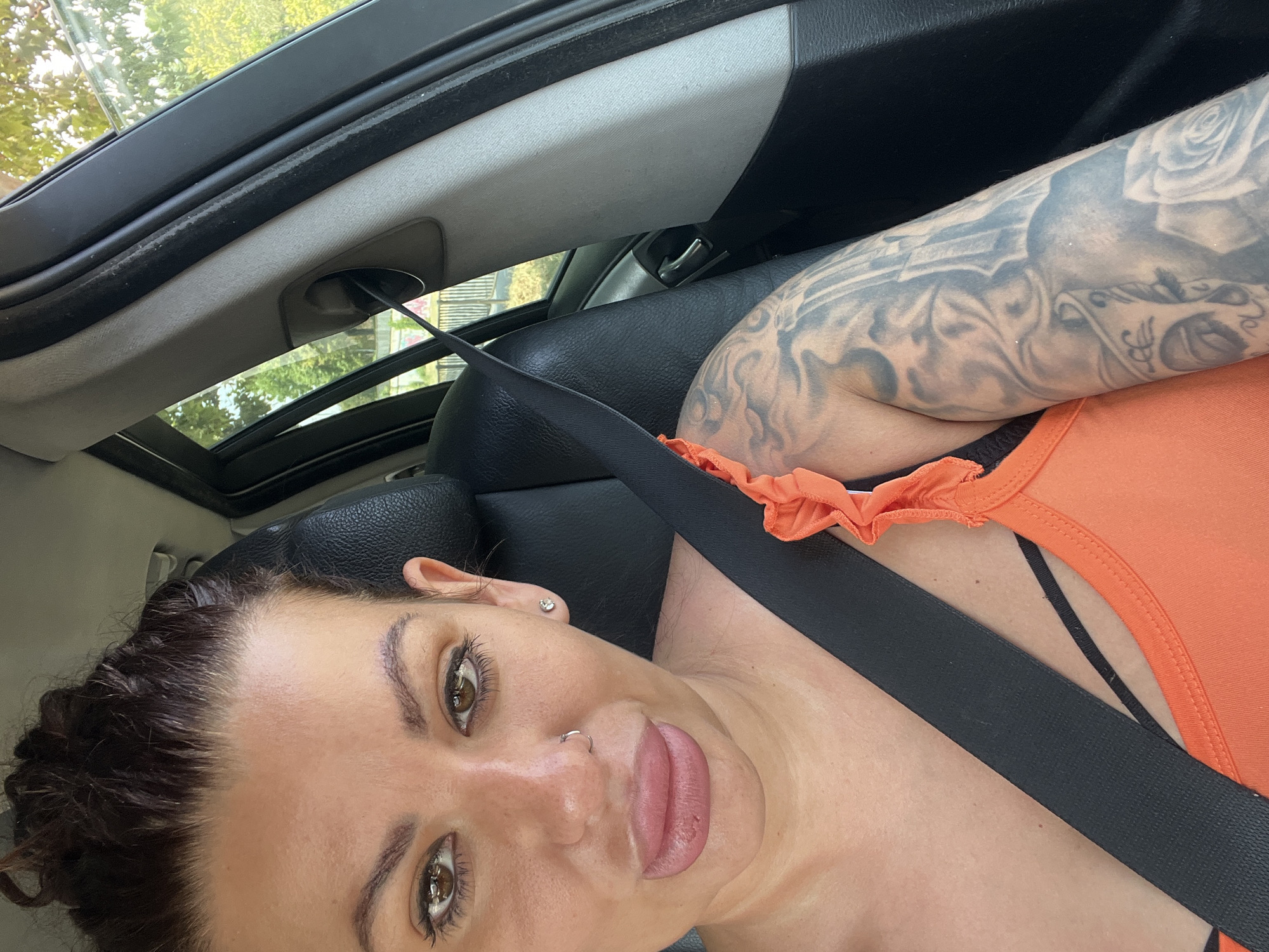 A woman with her hair in a bun and brown eyes is in the driver's seat of a car, looking forward at the camera. She has lip filler and is wearing an orange tank top with ruffled straps and a black bra. She has a seatbelt across her chest, and a large tattoo covers her left arm. The background shows the interior of the car and trees through the window, indicating an outdoor setting. The location is not specified in the city of Plovdiv, Bulgaria.