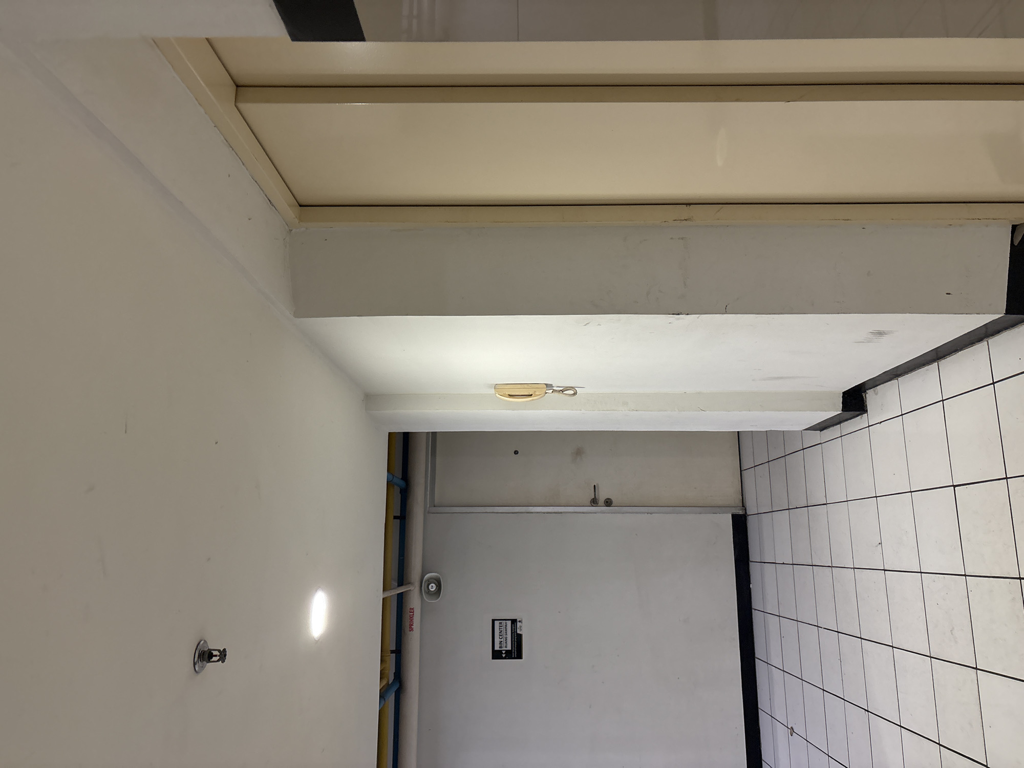 The image depicts a hallway with tiled floors and white walls. On the left wall, a sign reads "BIN CENTER RUANG SAMPAH". A white door with a silver handle is visible on the left. A sprinkler head is mounted on the white ceiling. To the right, a beige doorframe is present, with a portion of a beige door visible. A beige telephone handset is attached to the wall on the right. The environment appears to be an indoor corridor or hallway, possibly in a building in North Jakarta, Indonesia.