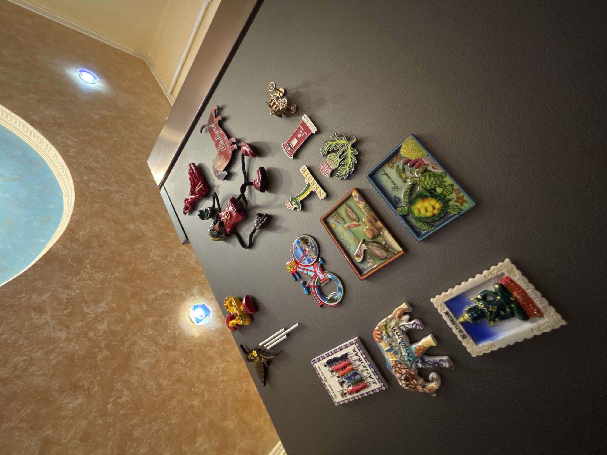 The image shows a refrigerator door covered in magnets. The magnets depict various locations and objects, including:
* "Budapest"
* "NESSIE SCOTLAND"
* "Barbados"
* "St Kitts & Nevis WEST INDIES"
* "Belek"
* "ZANZIBAR Hakuna Matata"
* "Benidorm"
* "Bangkok"
Other magnets are images of a bike, turtles, a bird with long beak, and other unidentifiable images. The refrigerator is located in an interior space. Part of the ceiling with a blue decorative element and walls of beige color is visible. The location in Odesa, Ukraine is unknown.