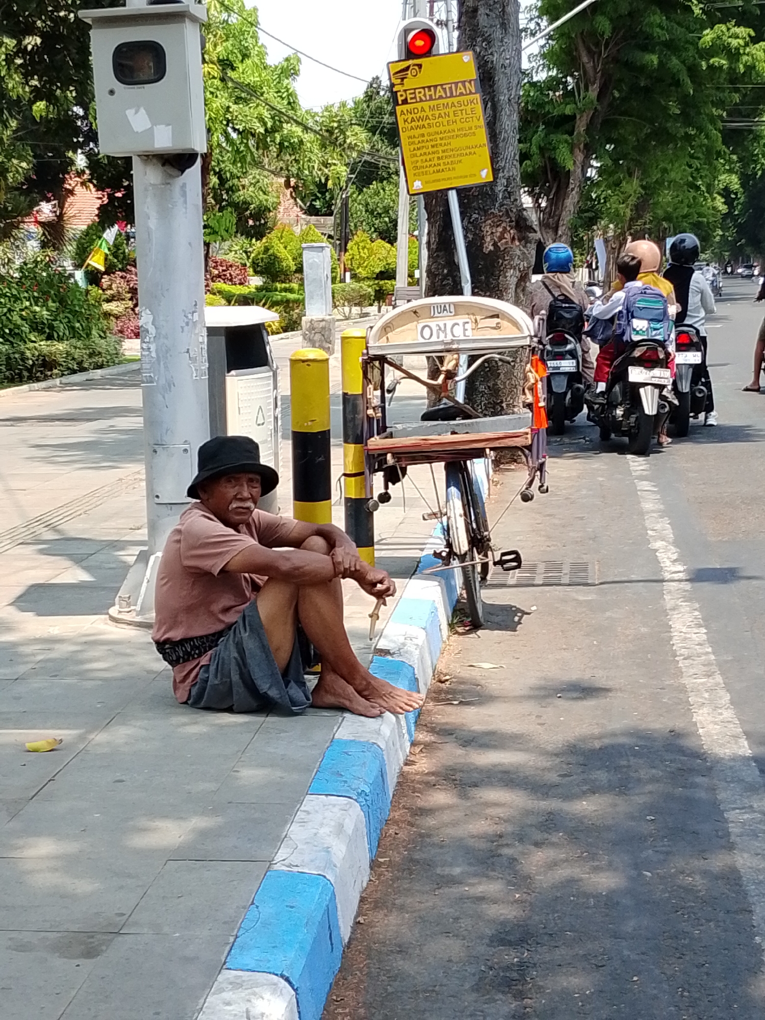 An elderly man with a black hat and a light pink shirt and gray shorts sits on the sidewalk in the city of Pasuruan, Indonesia. He is barefoot and appears to be holding a cigarette. A cart with a wooden top and bicycle wheels is parked behind him. In the background, several people on motorcycles are visible, some wearing helmets. A yellow sign with black writing and a red traffic light is attached to a pole nearby. The sidewalk is bordered by a curb painted with alternating blue and white stripes. The street is paved with asphalt. Trees and foliage line the background, suggesting a residential or commercial area.