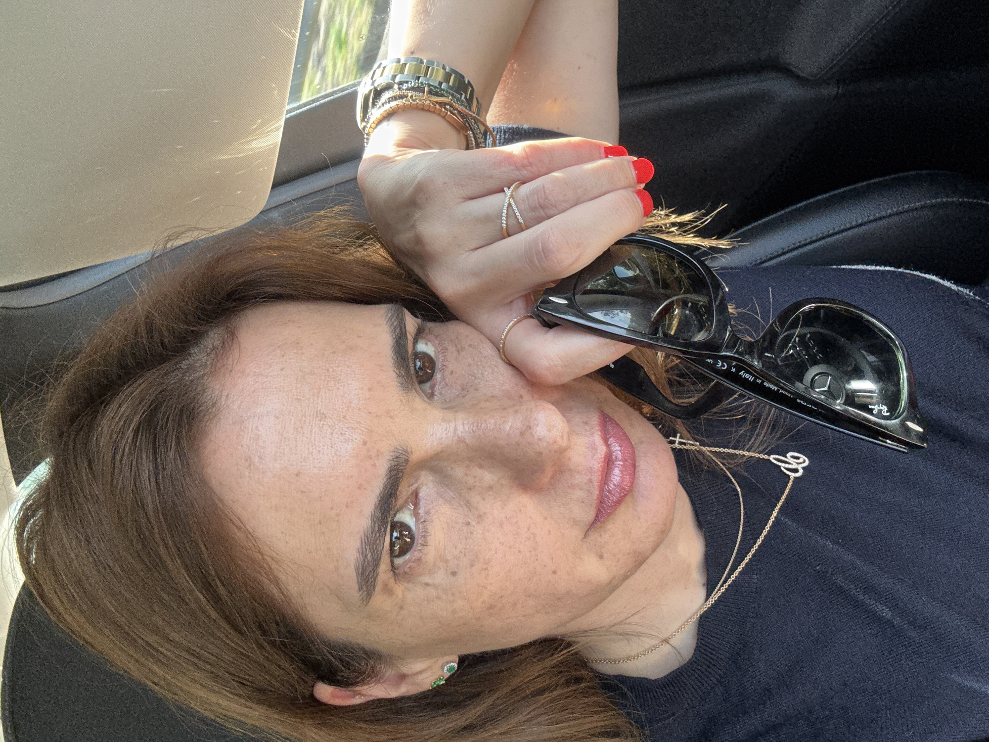 The image is a close-up shot of a woman in what appears to be a car. She is positioned with her head tilted, looking directly at the camera. The woman has light brown hair, freckles, and brown eyes. She is wearing a gold necklace and earrings, as well as several rings on her fingers. Her fingernails are painted red. She is holding a pair of black sunglasses. The background shows a car interior with the sun visor and a glimpse of foliage outside the window. It is not possible to confirm the location in Albergaria-a-Velha, Portugal, from this image alone.