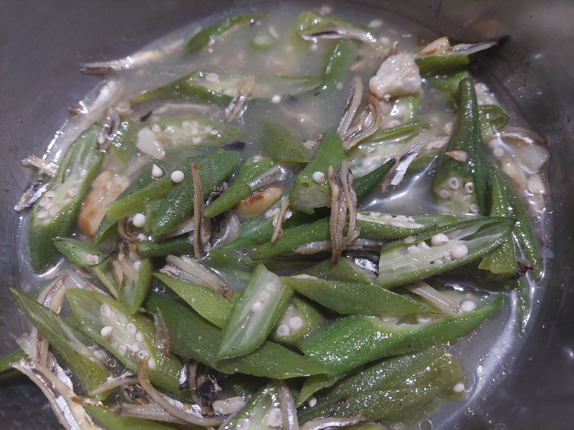 The image shows a close-up, overhead view of a dish containing sliced okra and small dried fish in a liquid. The okra is bright green and appears to be cooked, with visible seeds. The small fish are silvery and are mixed throughout the okra. The dish is in a metallic pot, and the lighting suggests an indoor setting. There are no people or other objects visible in the image. The context of Jelutung, Indonesia, is not directly inferable from the image itself.