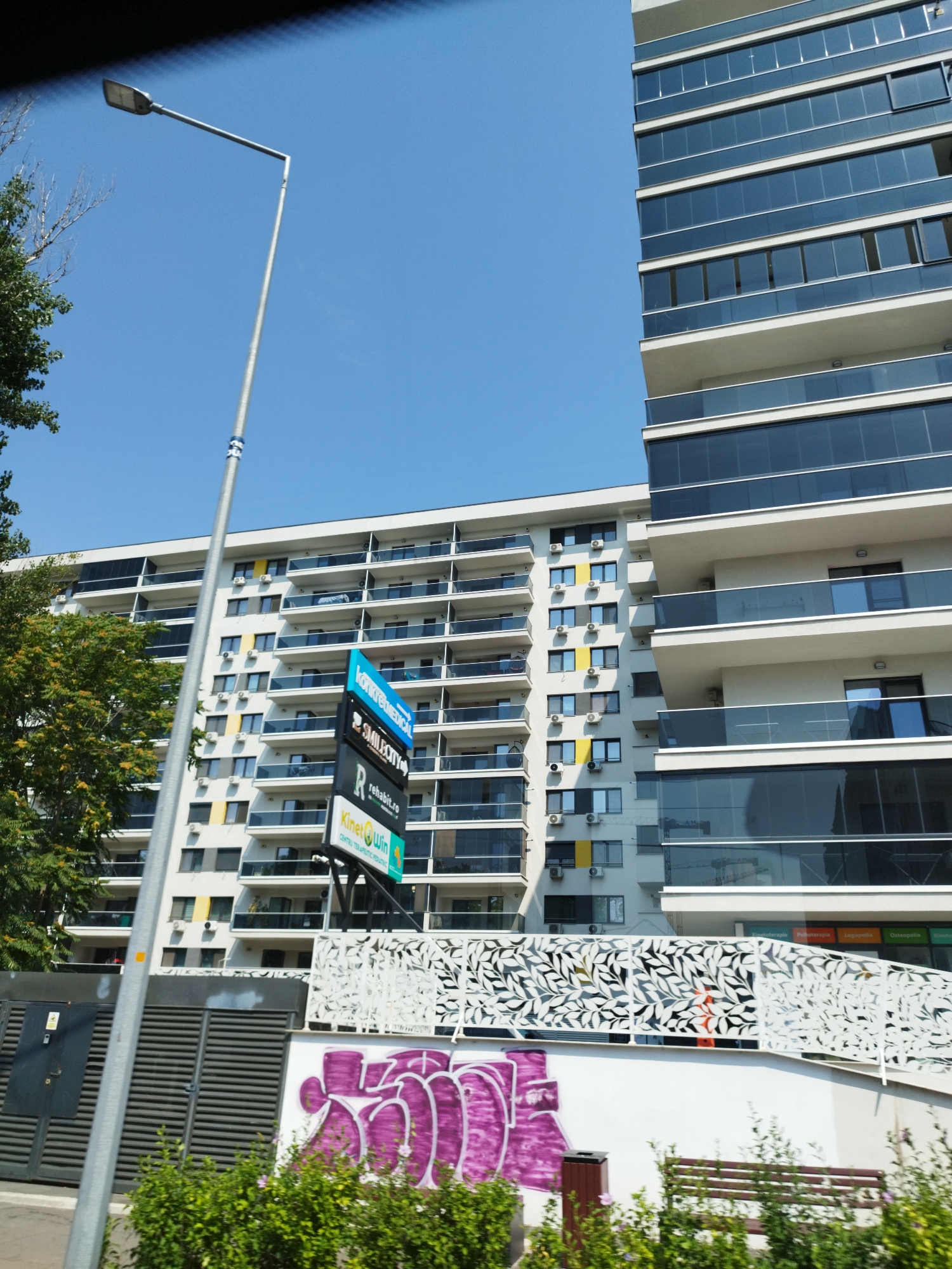 The image shows a modern building with a glass facade and balconies, adjacent to a multi-story apartment building with balconies and air conditioning units. A street lamp is visible in the foreground, partially obscuring a sign with various business names. There is graffiti on a wall in the lower right of the image, and dense green foliage. The sky is clear and blue. The buildings are located in Bucharest, Romania.
