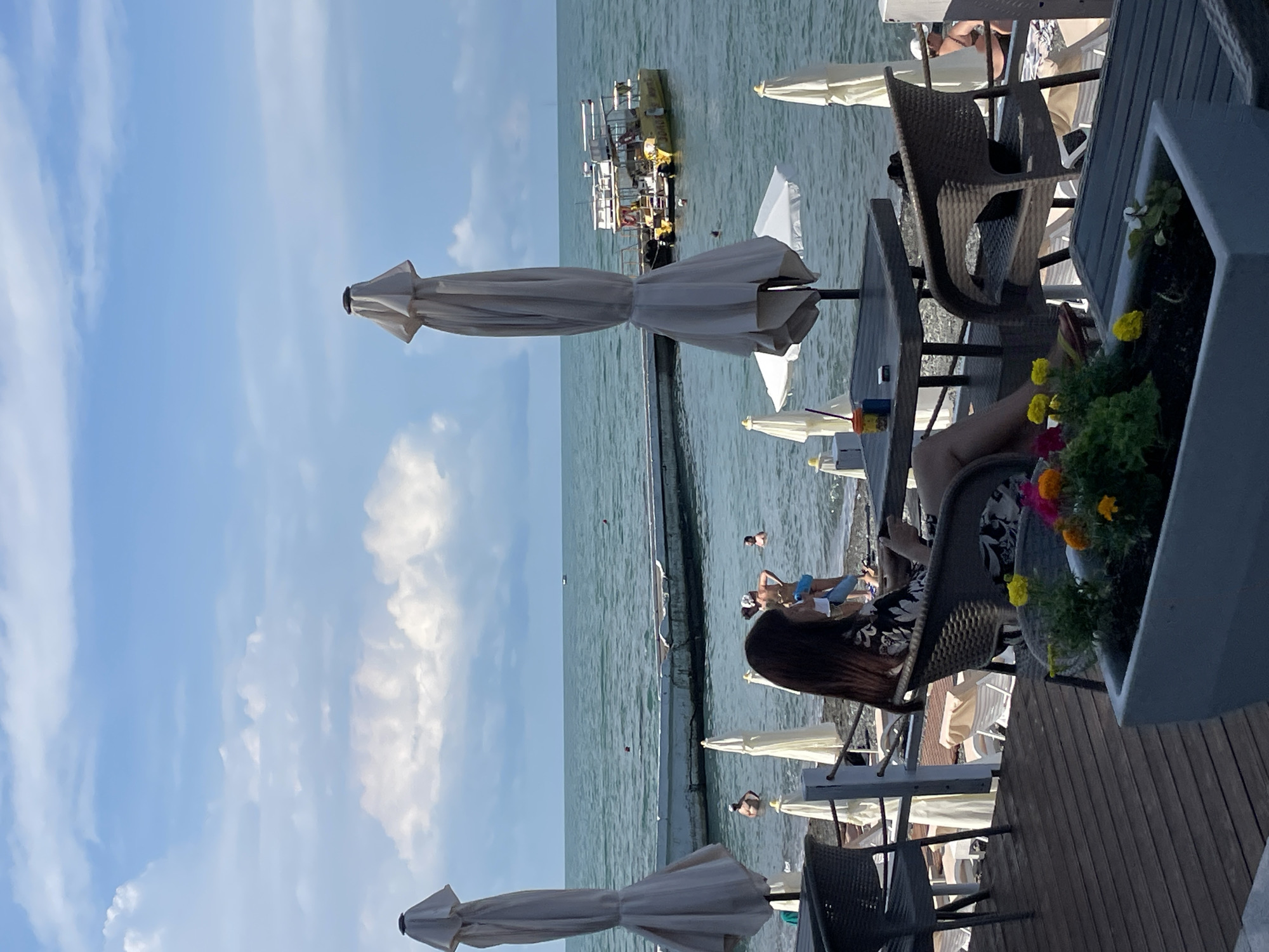 A woman with long dark hair, wearing a black and white floral print dress, sits in a wicker chair at a table on a wooden deck overlooking the Black Sea in Sochi, Russia. She is holding a smartphone in her right hand. In front of her, on the table, are two drinks, one with a red straw, and a pack of cigarettes. The scene is a beachside cafe with white umbrellas providing shade. Other beachgoers are visible in the water and on the shore, some swimming, others sunbathing. A yellow boat is docked near a pier in the distance. The sky is partly cloudy with a bright blue hue. A planter box with colorful flowers sits in the foreground.