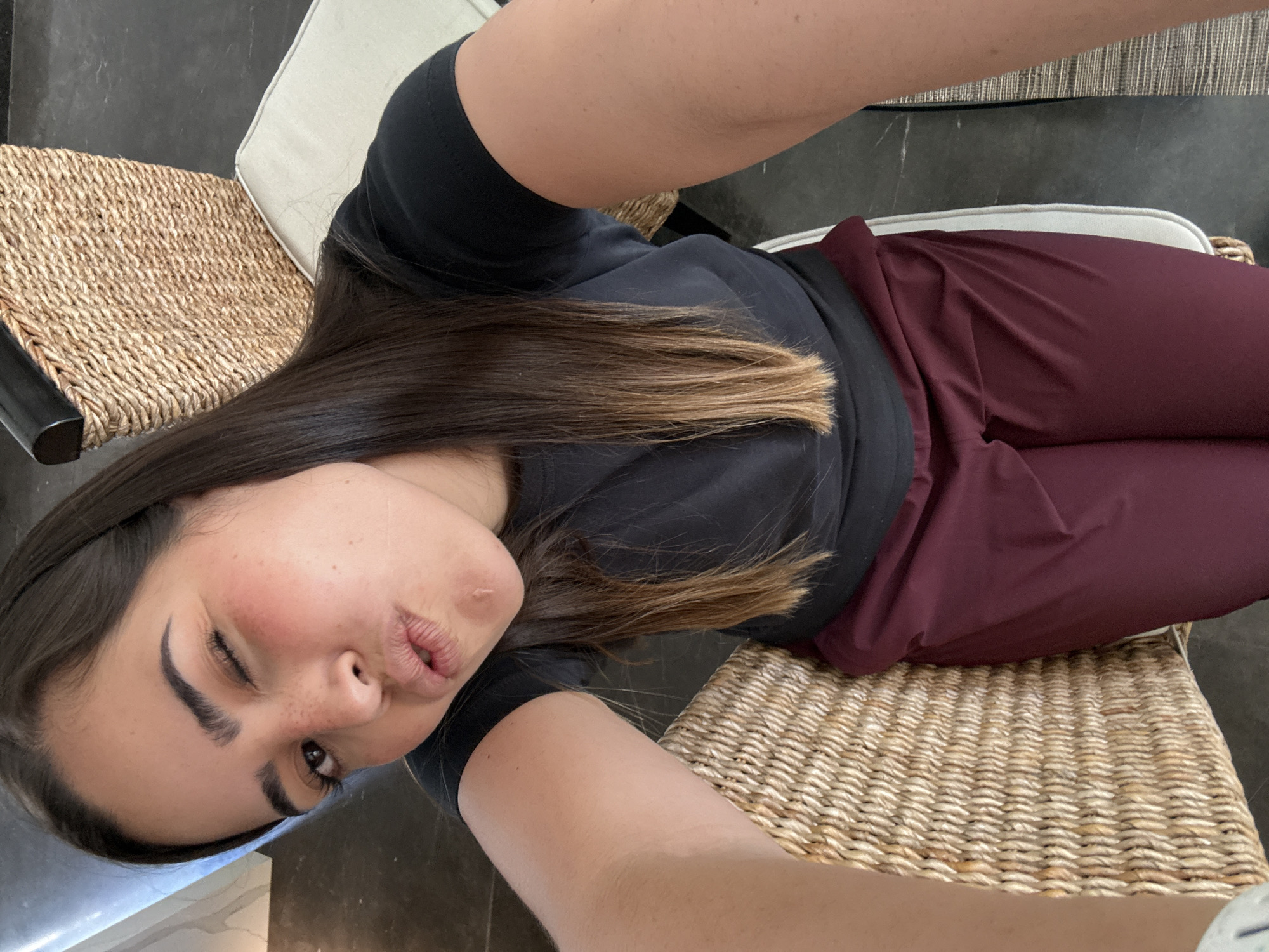 A woman is lying on her side on a chair or cushion, looking at the camera and puckering her lips. She is wearing a black t-shirt and maroon pants. The chair or cushion appears to be made of woven material. The floor beneath the chair is gray. The location is not discernible from the image.