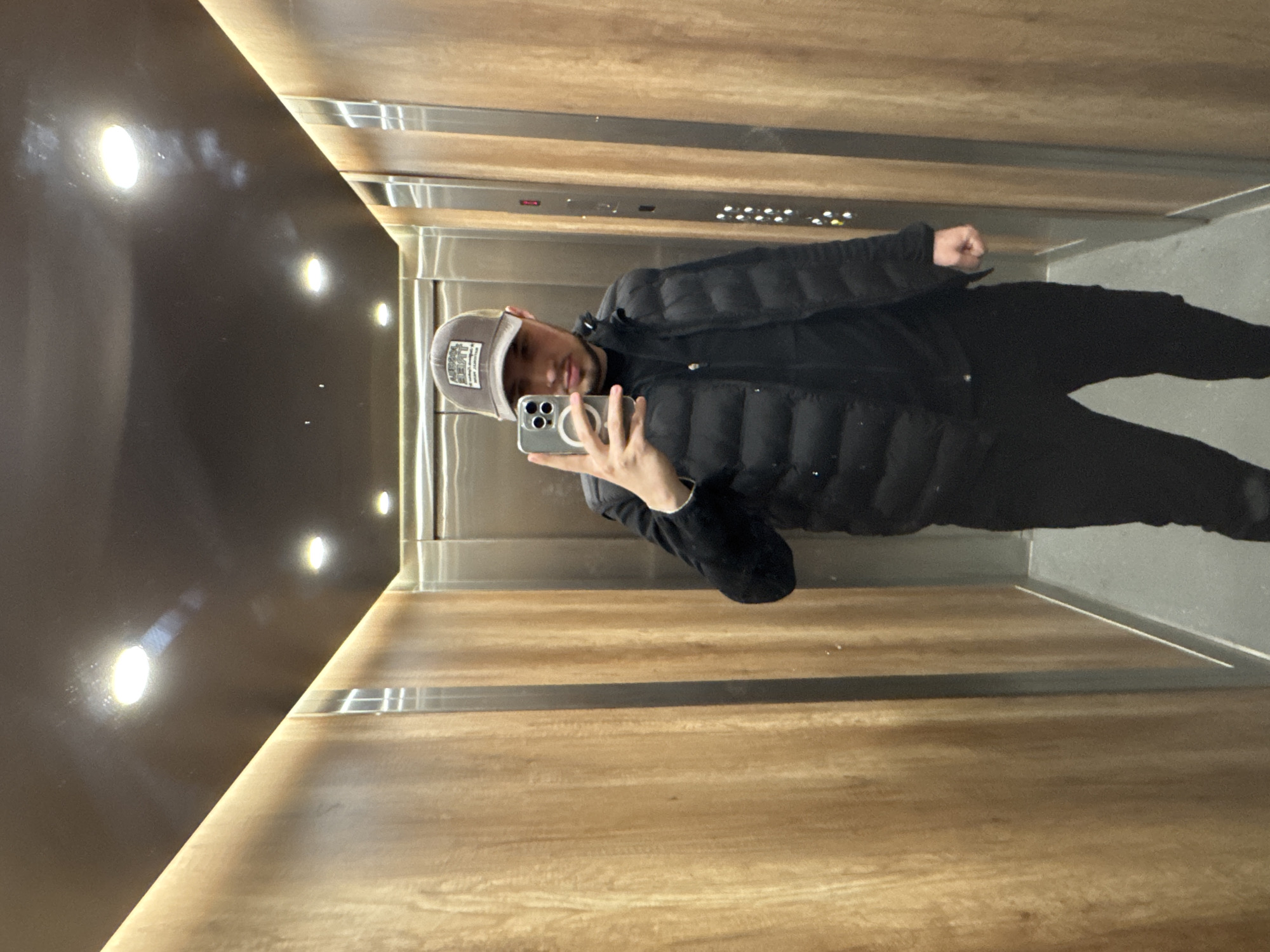 A young man is captured in a mirror selfie taken inside a modern elevator, situated in Sofia, Bulgaria. The image is rotated 90 degrees clockwise from a standard upright orientation.
**Subjects:** The primary subject is a young male, seen from the waist up, appearing to be taking his own reflection with a smartphone. He is wearing a dark, possibly black, puffer jacket, black trousers, and a grey baseball cap.
**Objects:** He holds a modern smartphone, likely an iPhone due to its distinct camera module, in his right hand. The elevator interior features sleek wooden paneling on the side walls and a dark, possibly metallic or reflective, ceiling/upper wall. Circular, warm-toned lights are visible on the ceiling and reflect on the dark surface. A narrow strip of ambient light runs along the join between the dark upper wall and the wooden paneling. A metallic elevator control panel with multiple buttons is partially visible behind him.
**Scene/Setting:** The scene is the interior of an elevator. The combination of warm wooden panels, modern lighting, and metallic accents suggests a contemporary and well-maintained building, possibly an apartment complex, office, or hotel in Sofia.
**Activity or event taking place:** The man is engaged in taking a selfie using his phone, capturing his reflection in the shiny surface of the elevator.
**Notable details:** The man's facial expression is neutral, looking directly at the camera. The lighting is artificial and bright, typical of indoor settings. The original image's rotation suggests a casual, perhaps impromptu, moment.
**Visible Text:** On the baseball cap, there is a rectangular patch with light-colored text, including "LIVEE" prominently visible, possibly a brand name or slogan, along with other smaller, indistinct text or a logo. No other text is clearly legible, though buttons on the elevator control panel have numbers and symbols.