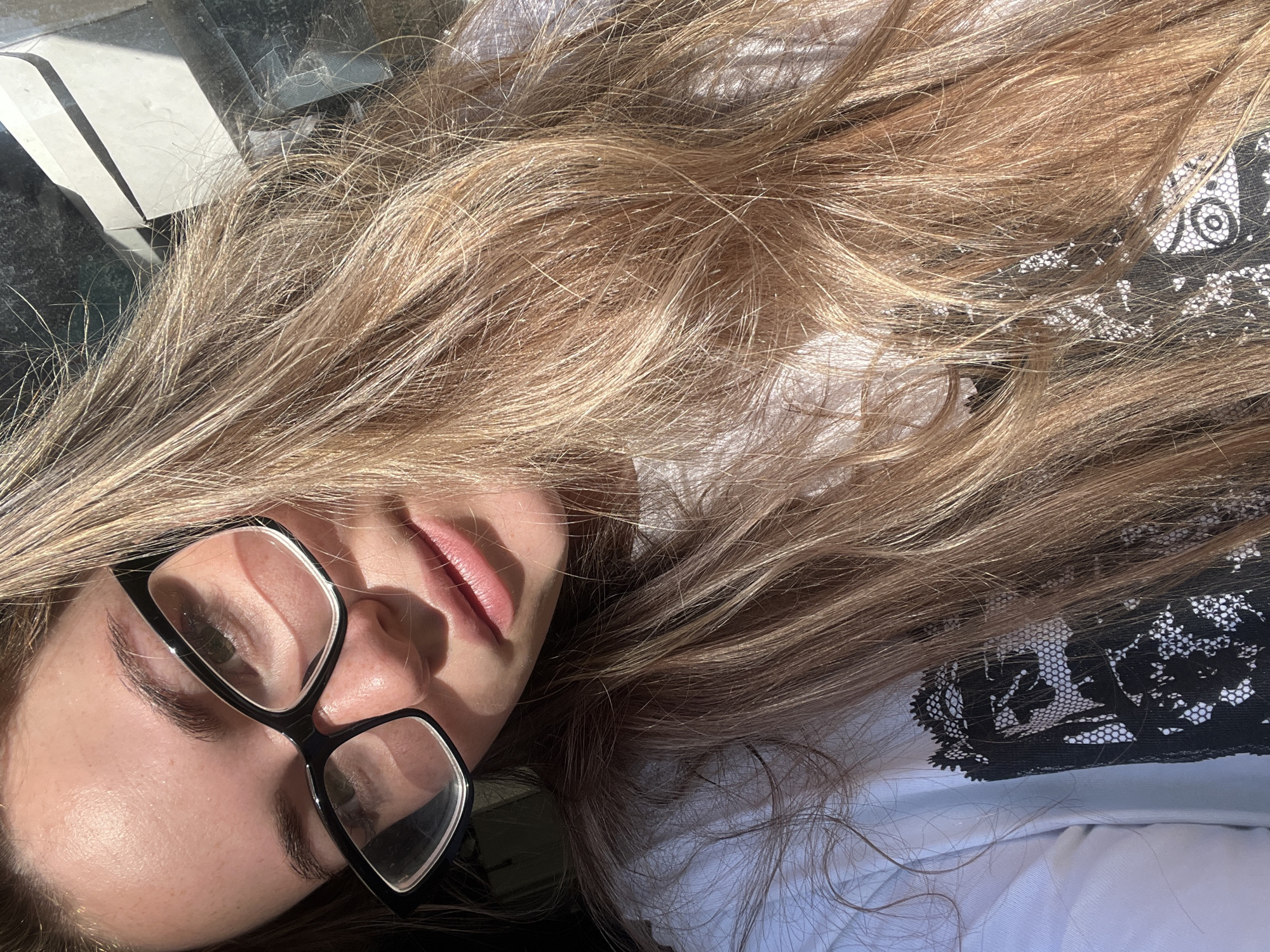 A woman with long blonde hair and black-framed glasses is lying down. Her face is partially illuminated by sunlight. She is wearing a white shirt with a black lace pattern. The location cannot be determined to be Chaikovsky, Russia based solely on the image.
