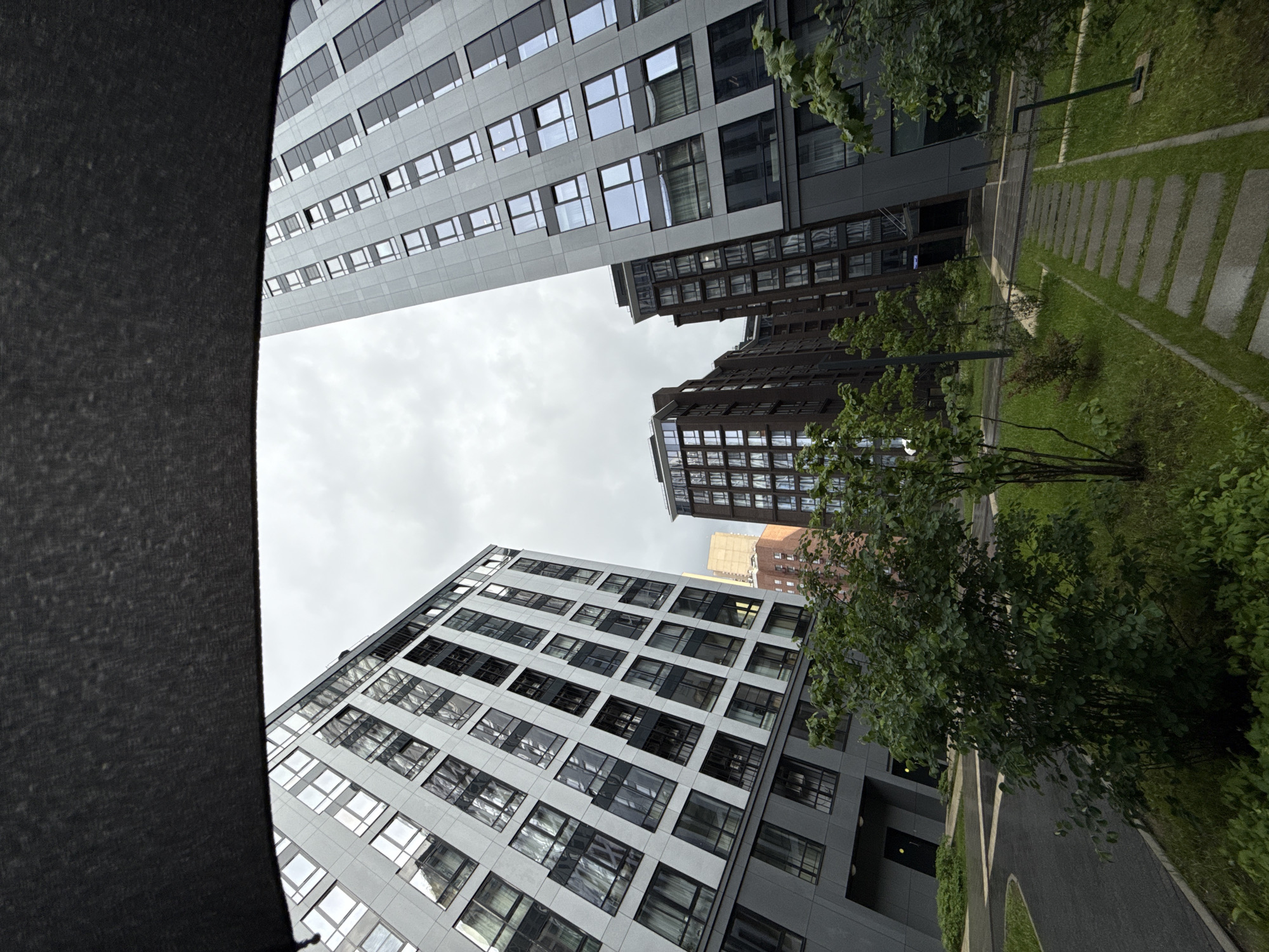 The image shows a low-angle view of several modern buildings in Moscow, Russia. The buildings are predominantly high-rise structures with repetitive window patterns, constructed with gray and brown materials. A green, landscaped area with trees and a stone-paved walkway is present on the right side of the frame. The sky is overcast, casting a muted light on the scene. A dark, blurry object occupies the left side of the frame, partially obscuring the view. No people are visible in the image.
