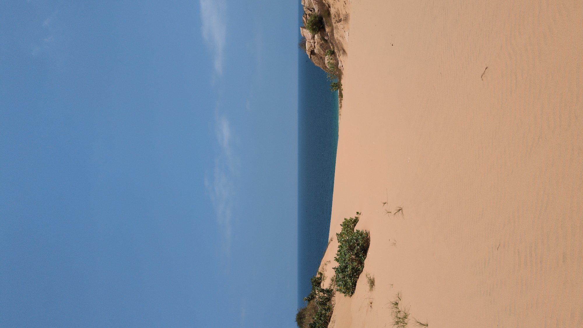 The image shows a section of the Mui Dinh sand dunes in Phuoc Dinh, Vietnam. The composition is vertically oriented, with the sky taking up roughly half of the left side. The sky is light blue with a few faint clouds. The right side depicts the sandy terrain, with varying textures and patterns indicating wind and erosion. The sand is a light beige color. Along the edge separating the sand from the sky, there is a strip of dark blue water. Patches of sparse vegetation, including green shrubs and some grasses, are visible on the sand dune. Some rocky formations can be seen atop the dunes. The image is a wide shot, showing the natural landscape.