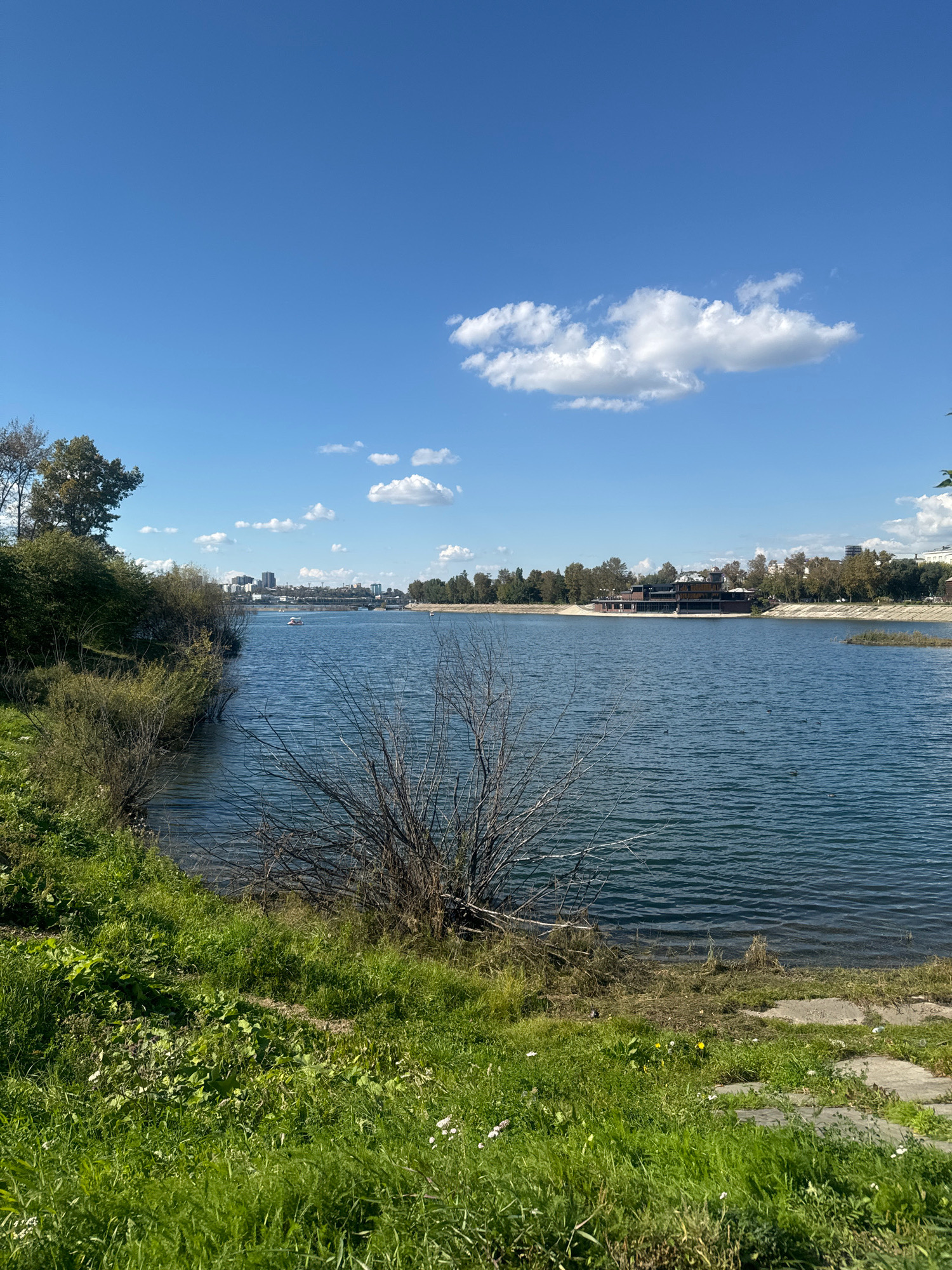 The image shows a wide, calm body of water, likely a lake or river, under a clear blue sky with scattered white clouds. The foreground features a grassy embankment with some bare, thorny bushes. In the middle ground, the water reflects the sky and surrounding trees, with a building complex situated on the far bank, appearing to be a restaurant or recreational facility. Several small boats, possibly pedal boats, are visible on the water. In the distance, a city skyline with buildings is discernible. The scene suggests a park or recreational area in a city, possibly during the daytime on a pleasant day. The specific location in Irkutsk, Russia, cannot be determined from the visual information alone.