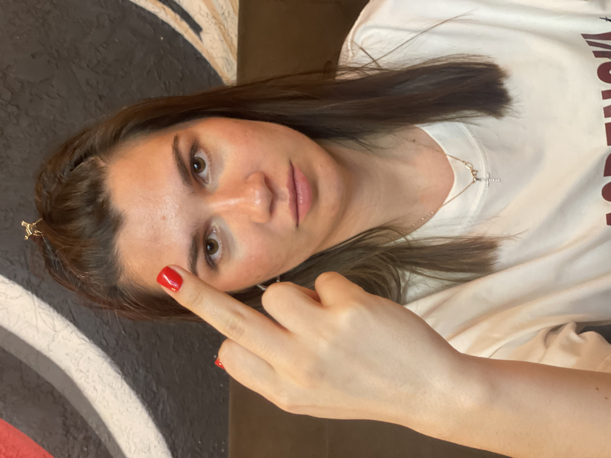 A woman is indoors, facing the camera and extending her middle finger with a red manicure. She has brown hair and brown eyes, wearing a white t-shirt and a silver necklace with a cross pendant. The background includes a circular design with black, white, and red elements. Location information (Krasnoyarsk, Russia) cannot be confirmed by visual elements in the image.