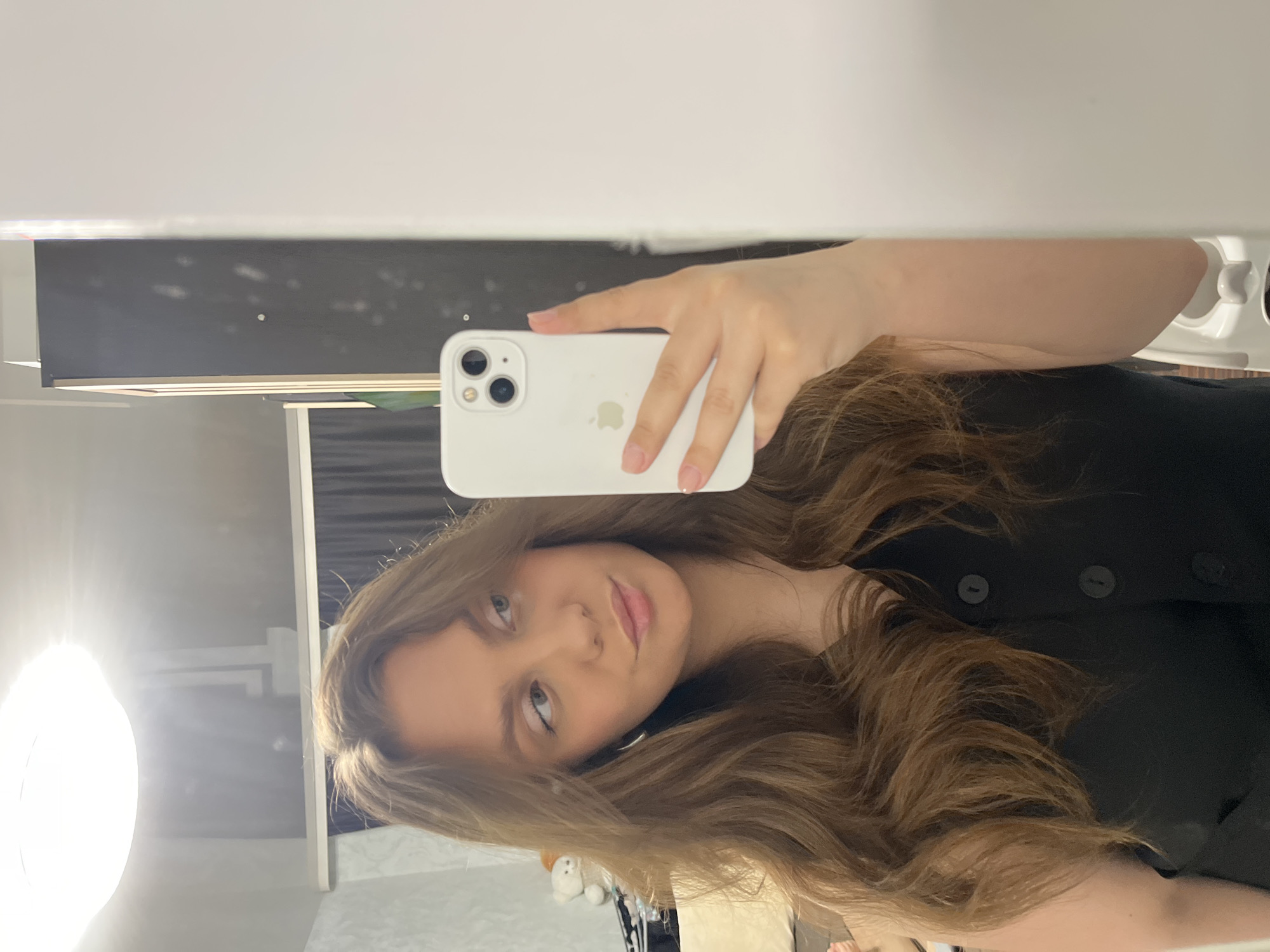 A young woman is taking a mirror selfie with a white iPhone. She is wearing a black top with buttons. Her hair is long, wavy, and light brown. The background includes a white wall, a dark gray shelf with some items on it, and an artificial ring light. The location appears to be inside a residential space, most likely in the city of Chaikovsky, Russia, but this is speculative based on the user's profile.