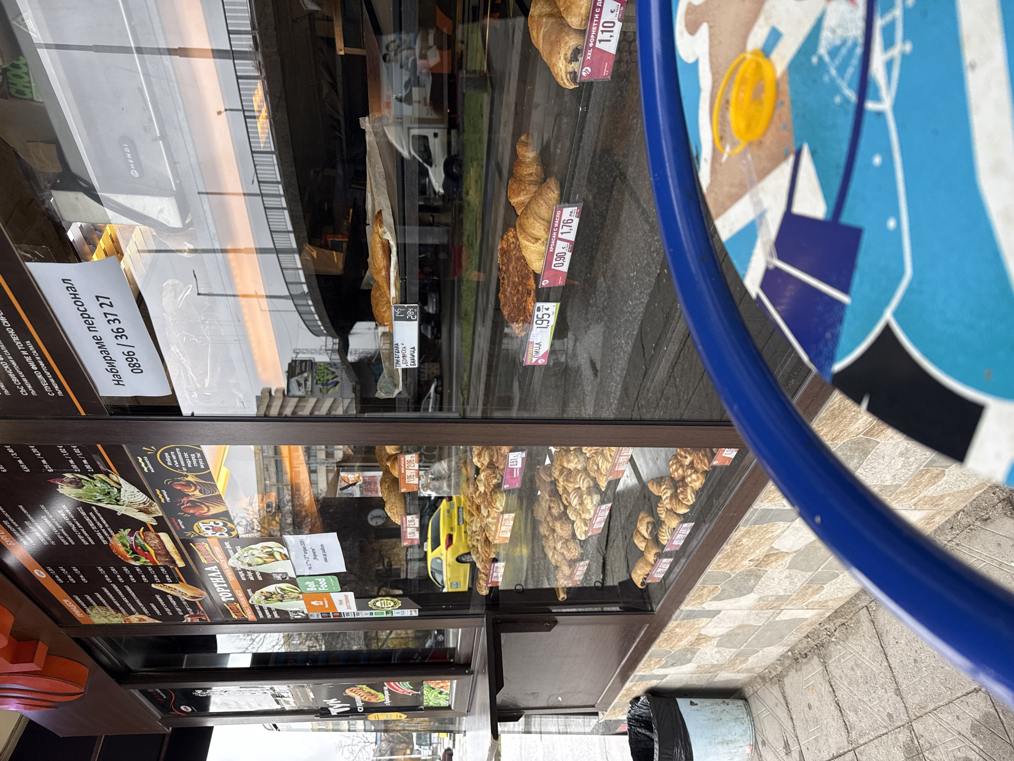 The image depicts the exterior of a bakery or fast-food establishment in Plovdiv, Bulgaria. A display case showcases various pastries like croissants and pizza slices. Pricing labels are visible in Bulgarian currency (лв) and Euros. A menu board advertises items like burgers, wraps, and salads, alongside delivery service information. The reflections in the glass show a street scene, with a parked yellow taxi and other vehicles. A blue-rimmed table with a design is visible in the foreground. A trash can is located nearby. A recruitment sign in Bulgarian advertises for staff at the establishment.