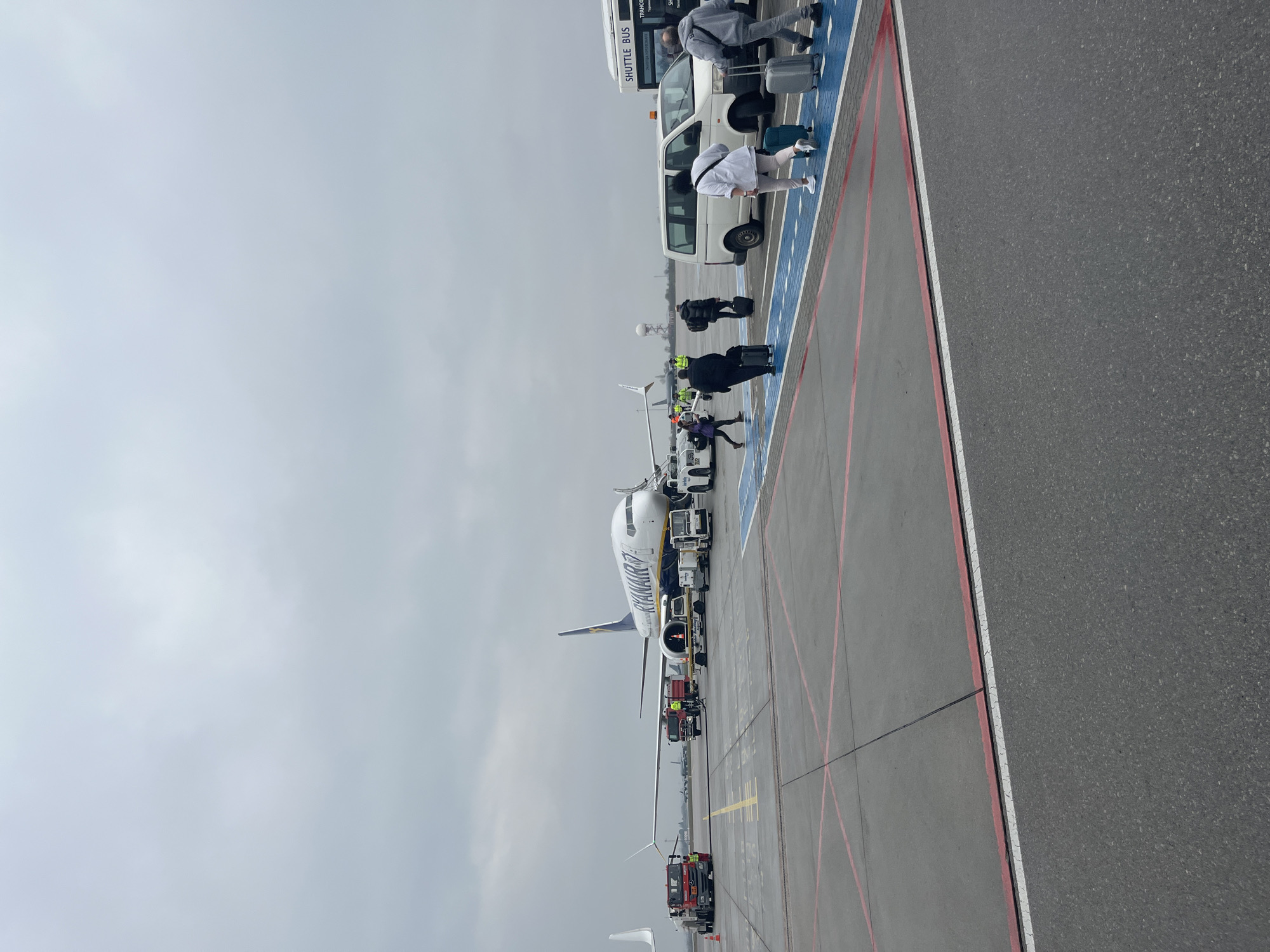 The image shows a Ryanair airplane at Sofia Airport in Bulgaria. Passengers are boarding or disembarking, some being transported by a shuttle bus. Ground support vehicles are also present near the aircraft. The sky is overcast, suggesting a cloudy day. The location is an airport apron with painted markings for vehicles and pedestrian routes.
