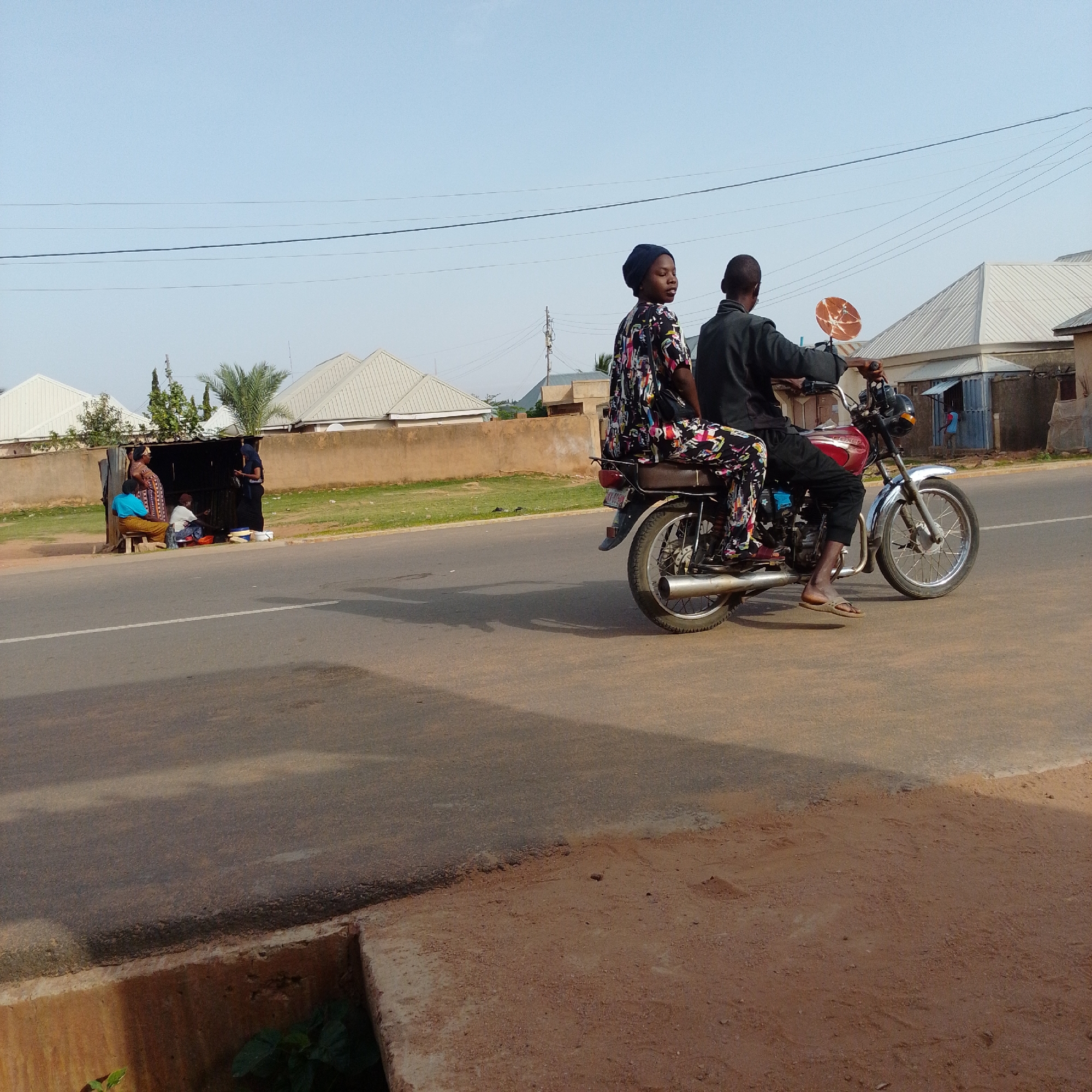 A photograph captures a daytime scene on a paved road in Gombe, Nigeria. The foreground features a segment of the road with a concrete drainage channel to the left. In the midground, two individuals are riding a red and silver motorcycle. The person in the front, wearing a dark long-sleeved shirt, is operating the motorcycle. The person behind, a woman, is wearing a colorful patterned dress and a dark head covering, and she is holding a circular, orange object, possibly a fan or a small plate. To the left of the road, near a cluster of buildings with white, angular roofs, a small open-fronted stall is visible. Three people are gathered at this stall: one woman in a blue top and patterned skirt sits on a wooden bench, another woman in a patterned dress stands, and a third person in white is seated behind the stall. The background shows more of the buildings and a clear sky with visible power lines.