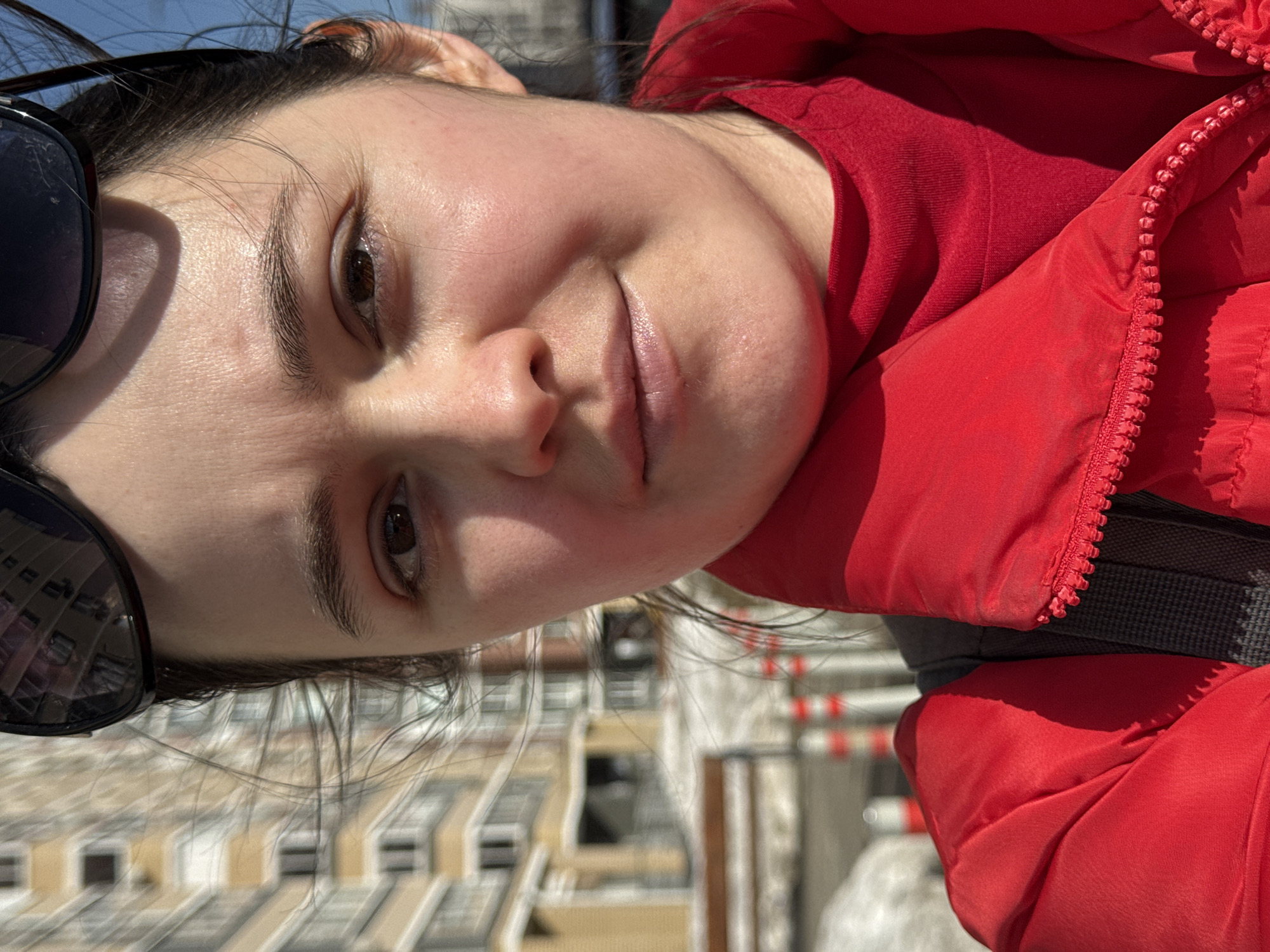 A selfie photograph depicts a light-skinned woman with dark hair wearing a red jacket and sunglasses. The background shows a city building, and based on architectural style, this may be Izhevsk, Russia. The woman is angled in the shot so she can be seen more clearly than the background building.