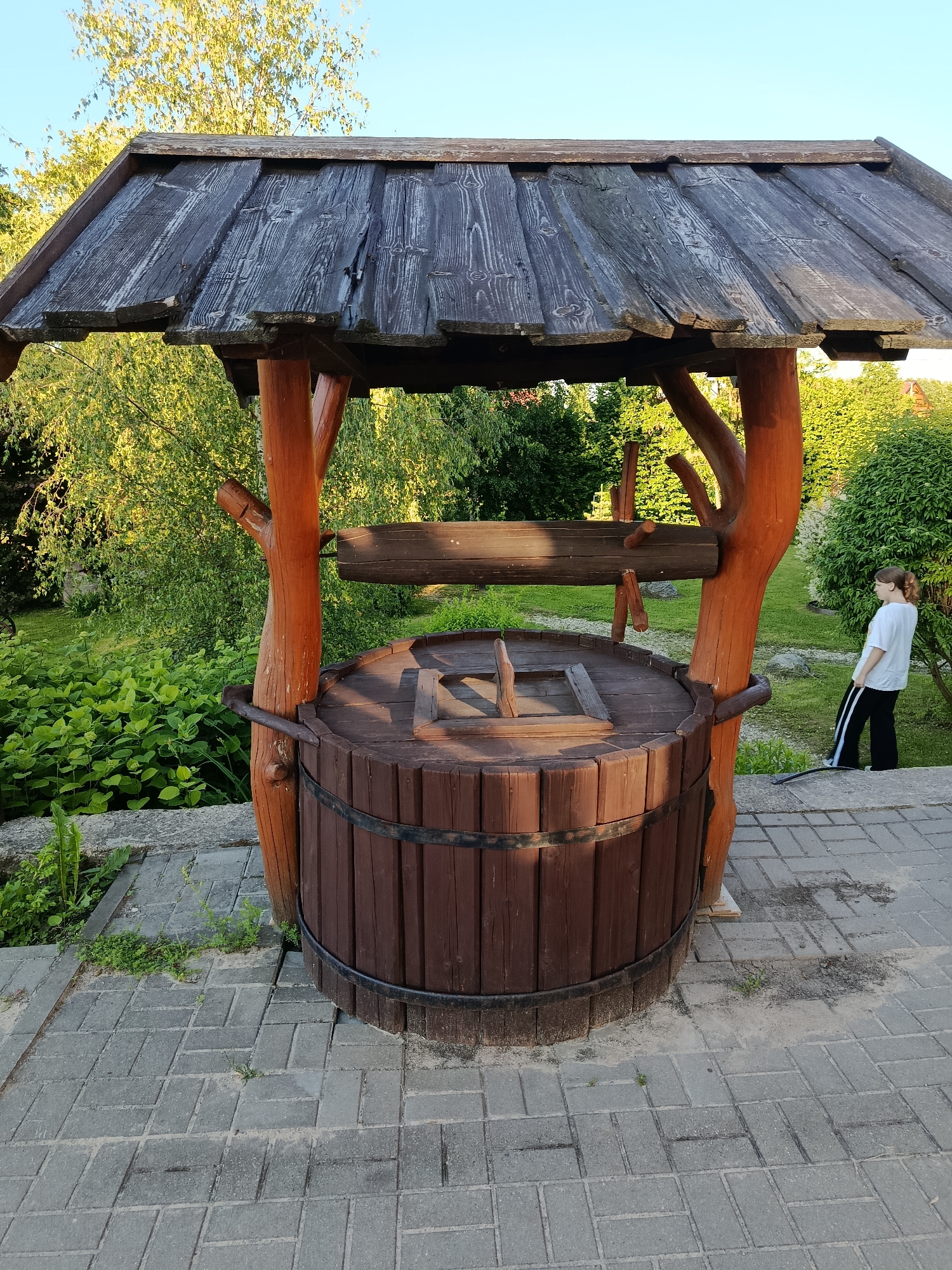 The image shows a rustic wooden well structure in an outdoor setting. The well has a large, barrel-shaped base made of vertical wooden planks held together by two dark metal bands. The well is covered by a slanted wooden roof supported by four thick, reddish-brown wooden posts. A wooden beam with two wooden cranks extends horizontally over the well opening.
To the right of the well, a young person with blonde hair tied in a ponytail is standing, wearing a white t-shirt and black pants with white stripes. They are looking towards the well structure.
The well is situated on a paved walkway made of grey interlocking bricks. Behind the well, there is lush green foliage, including bushes and trees. The background suggests a garden or park environment. The sky is clear and blue, indicating a daytime scene. The lighting suggests it is either late afternoon or early morning, with the sun casting shadows and highlighting the textures of the wood. The overall impression is one of a traditional or decorative well feature in a landscaped area. The image was likely taken in the Мяжанскі сельскі Савет, Belarus, as indicated by the context provided.