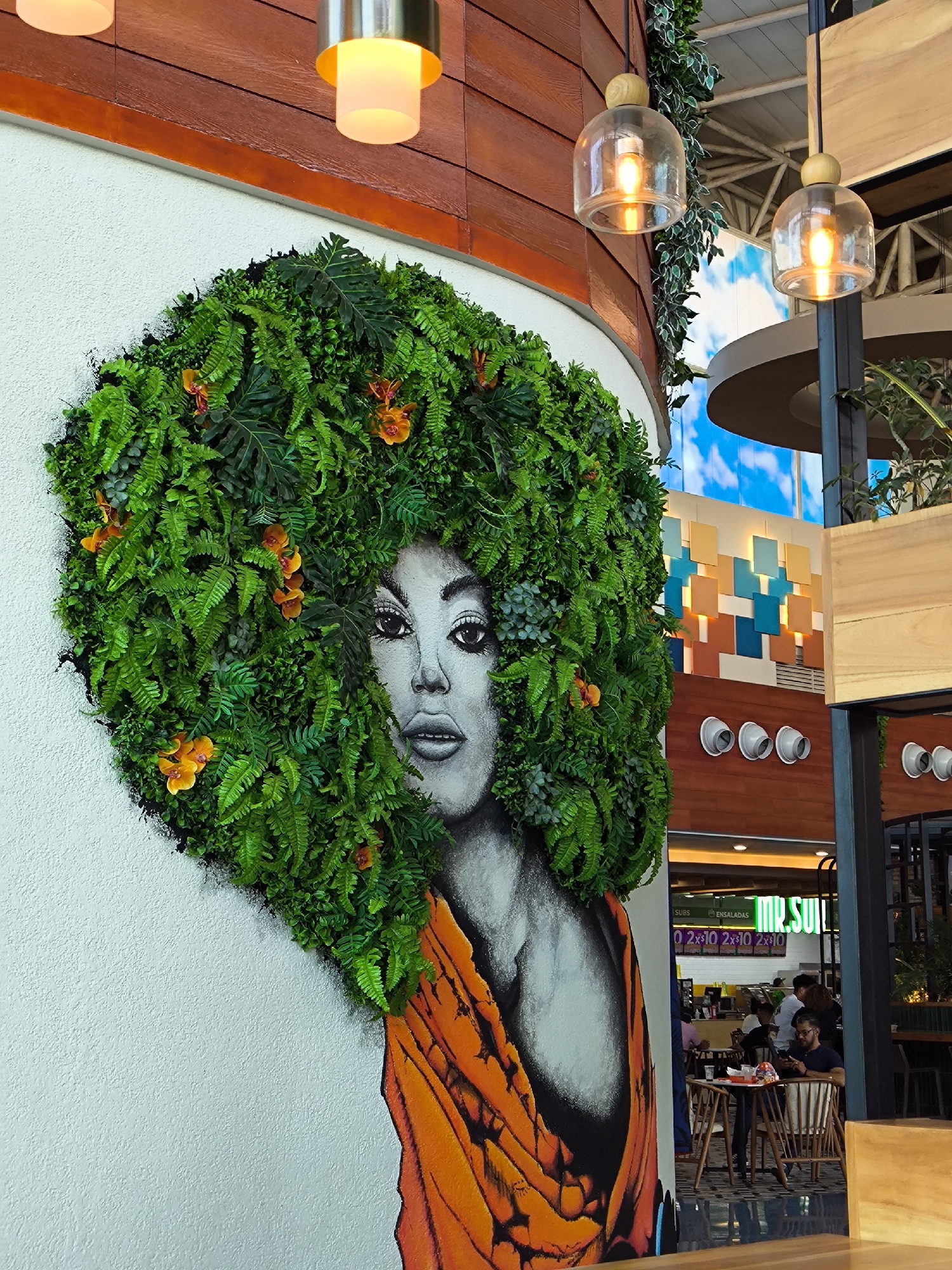 A wall mural depicting a woman with an afro made of green foliage and orange flowers is on a white wall. The woman has gray skin, a black chest, and an orange shawl. A lamp hangs from the ceiling above the mural. In the background, people sit at tables inside a restaurant called "Mr. Subs." The location is Porlamar, Venezuela.