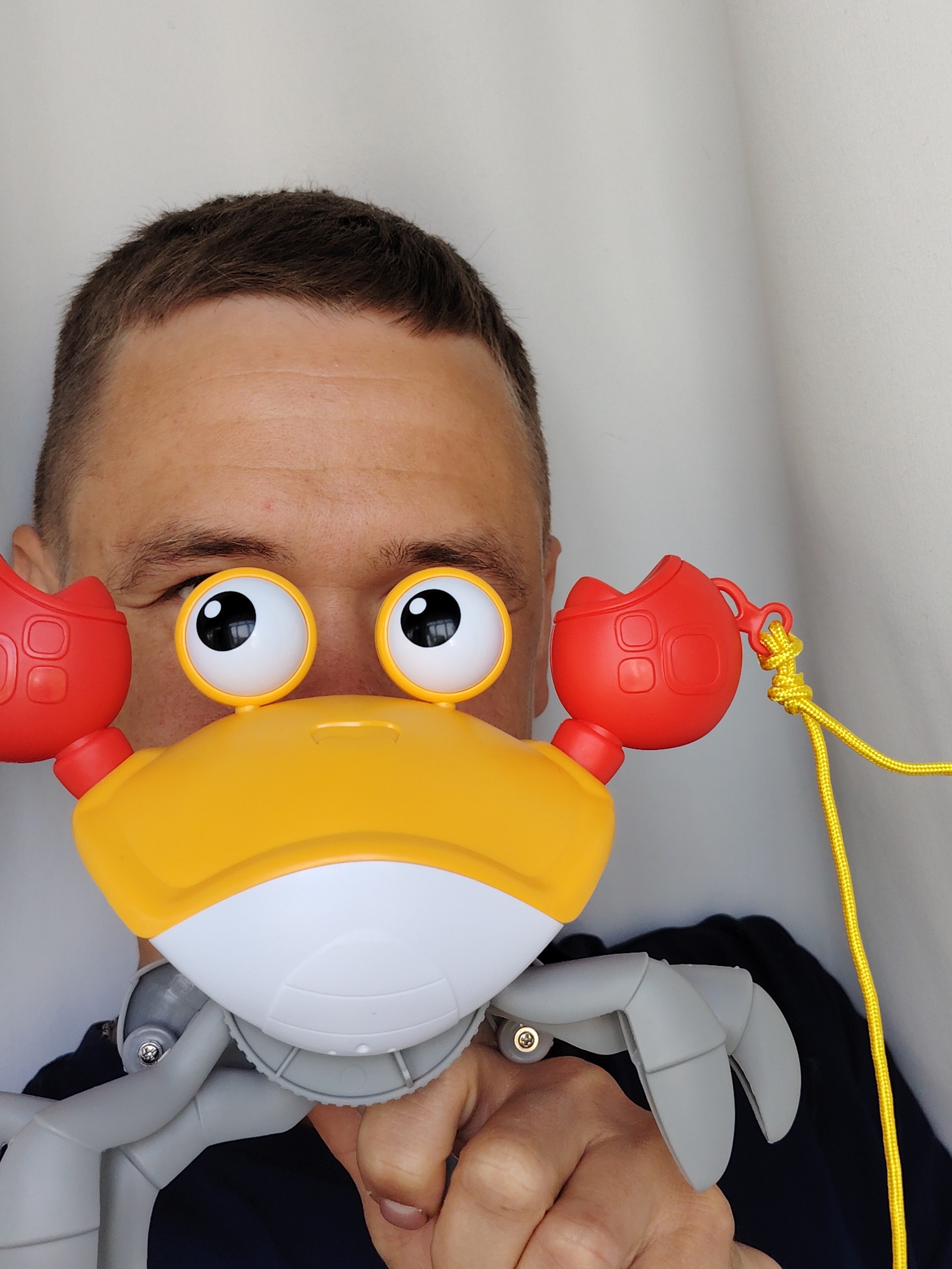 A person with short brown hair is holding a toy crab up to their face. The toy crab is yellow and white with red claws and googly eyes. The person is looking through the googly eyes of the crab. The background is a plain white wall. The crab also has gray legs. There is a yellow cord attached to the red claw of the crab. It is not possible to identify the location of the photo as Chaikovsky, Russia, or any other location based on the image alone.