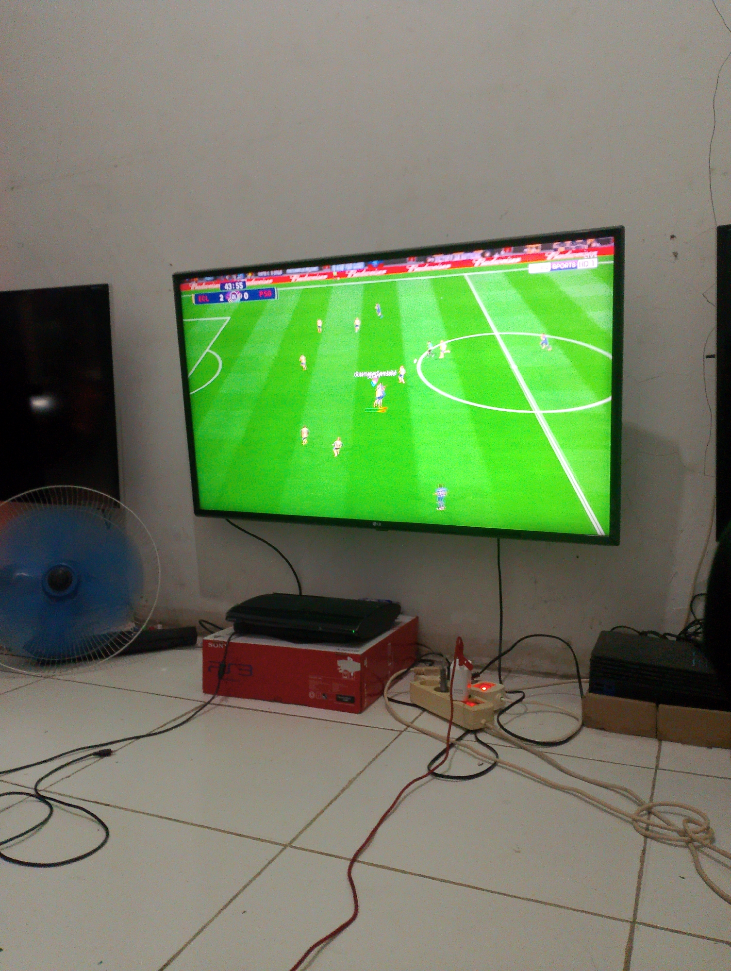 The image depicts a living room or entertainment space in Tangerang, Indonesia, with a large flat-screen television displaying a soccer match. The scoreboard shows "ECL 2 0 Psg" and the time is 43:55. The television is mounted on a wall, and below it, on the floor, are various electronic devices, including a PlayStation 3 console with its red box, a power strip with illuminated red lights indicating power, and several cables. A blue electric fan is positioned to the left of the television. The room has tiled flooring and plain white walls. The overall impression is one of a casual home entertainment setup.