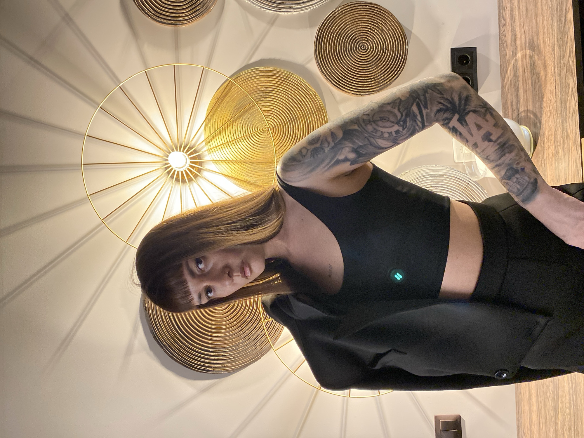 A medium shot of a woman with brown hair and bangs, wearing a black crop top and blazer, standing indoors against a wall decorated with circular golden ornamental pieces. The woman has a tattoo on her left arm, and appears to be looking directly at the camera. No information is provided to determine the location is in the city of Votkinsk, Russia.