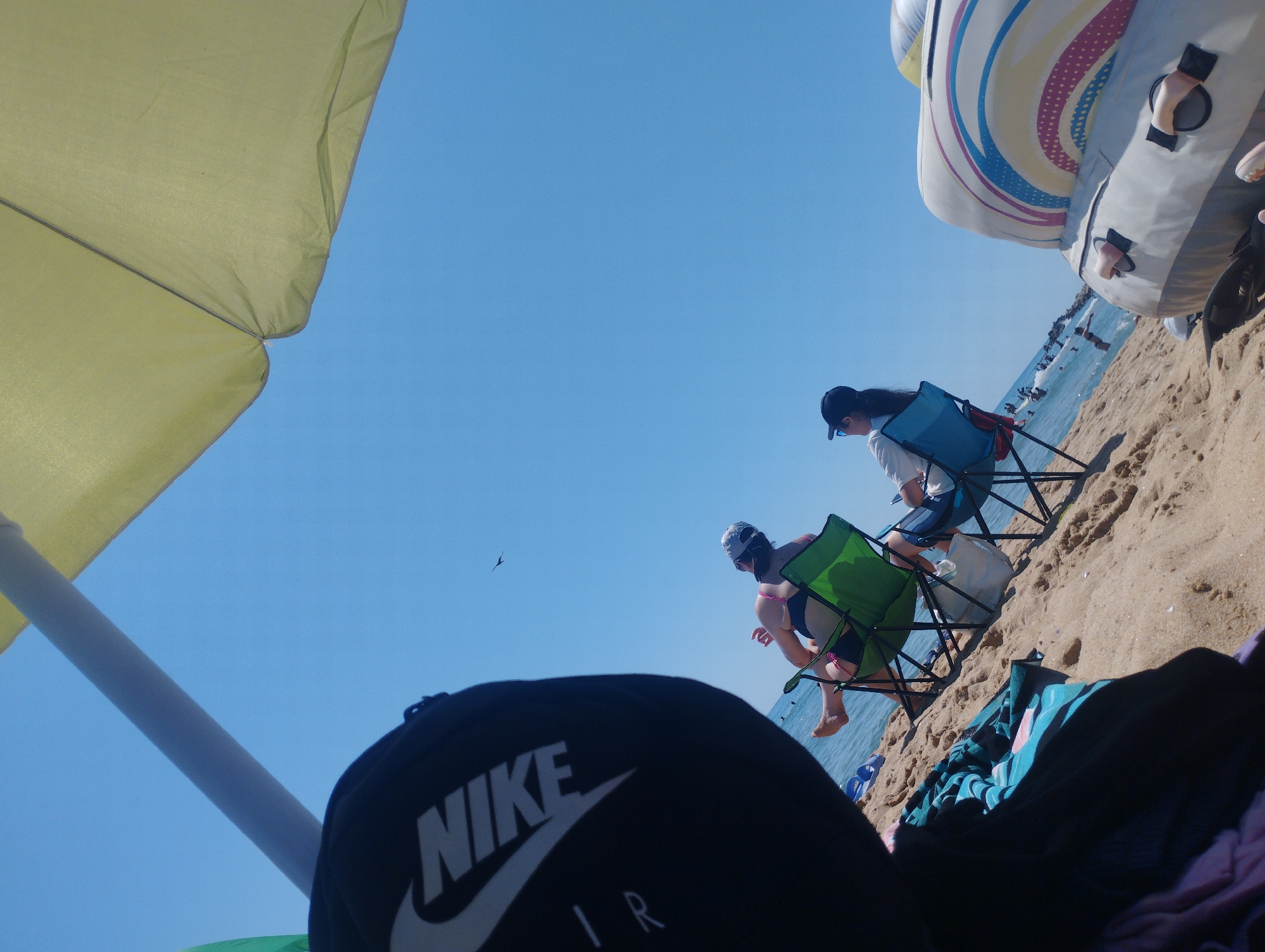 An outdoor, ground-level, wide-angle shot captures a beach scene on a sunny day. A yellow beach umbrella dominates the left side of the frame, angled upwards. In the background, the ocean meets the sandy shore. Two people are seated on beach chairs, facing away from the camera towards the water. The person on the right is wearing a blue swimsuit and a baseball cap, while the person on the left is wearing a white top and a baseball cap. A large inflatable toy with colorful swirling patterns is partially visible in the upper right quadrant. In the foreground, a black cap with a white Nike logo and "AIR" text is positioned at the bottom center. The sky is a clear, bright blue. The overall impression is a relaxed beach day.
