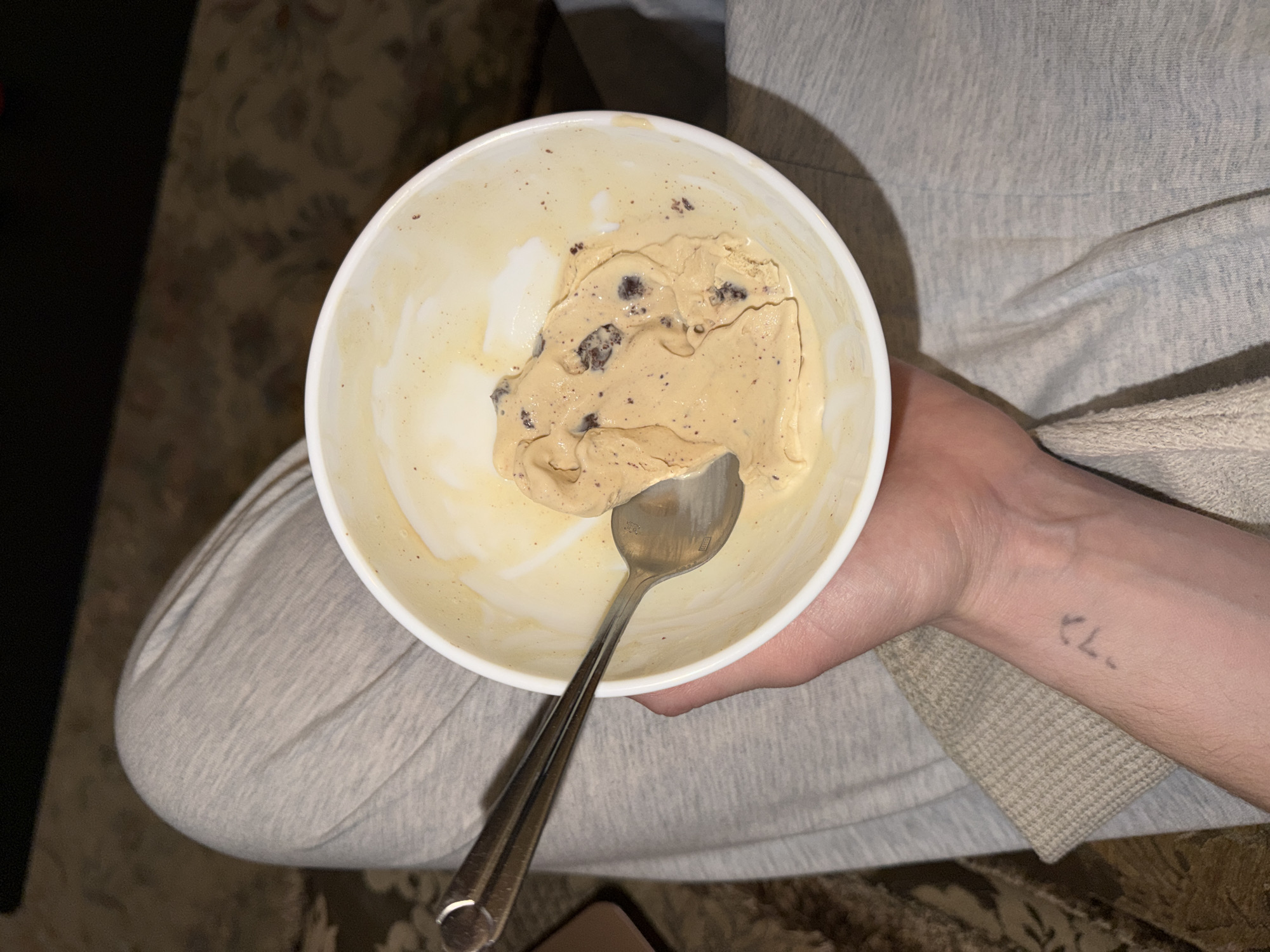 A person is holding a white bowl of partially eaten ice cream with chocolate chips. A silver spoon is inside the bowl. The person is wearing light gray sweatpants and a long-sleeved shirt. The background shows a patterned floor. There is no indication that this is in Mishutkino, Russia.